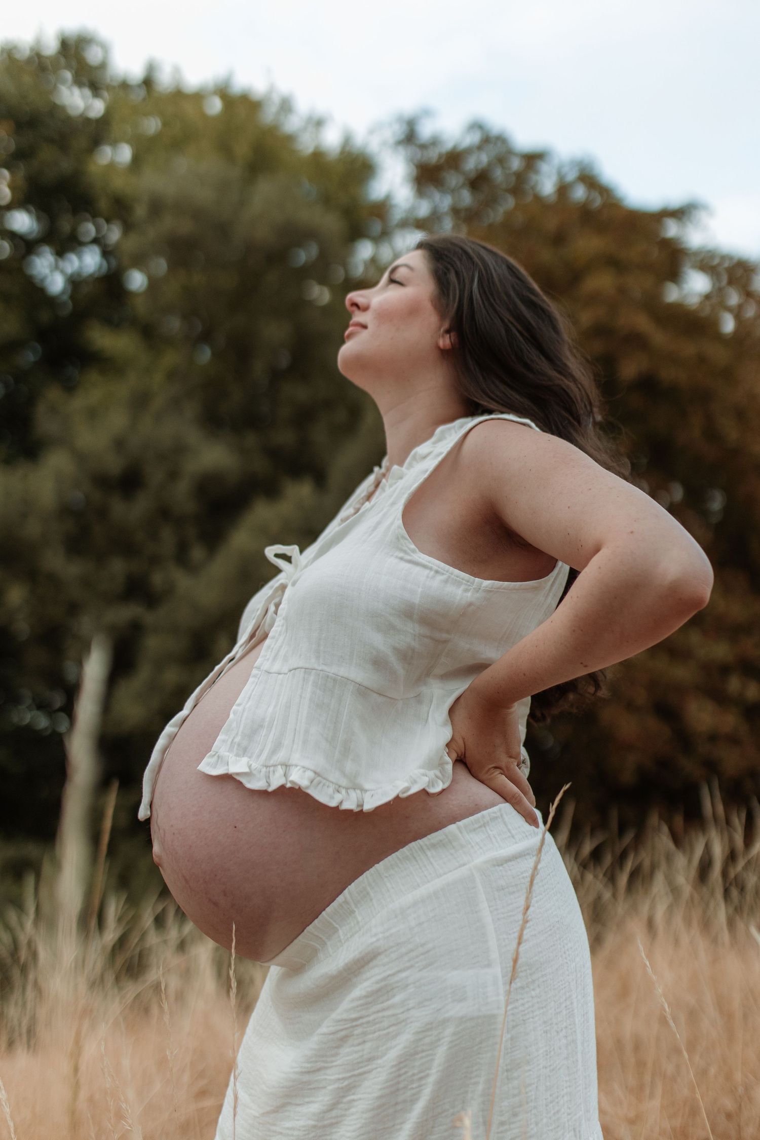 A pregnant woman in a white outfit standing in a golden field showing her bare bump.