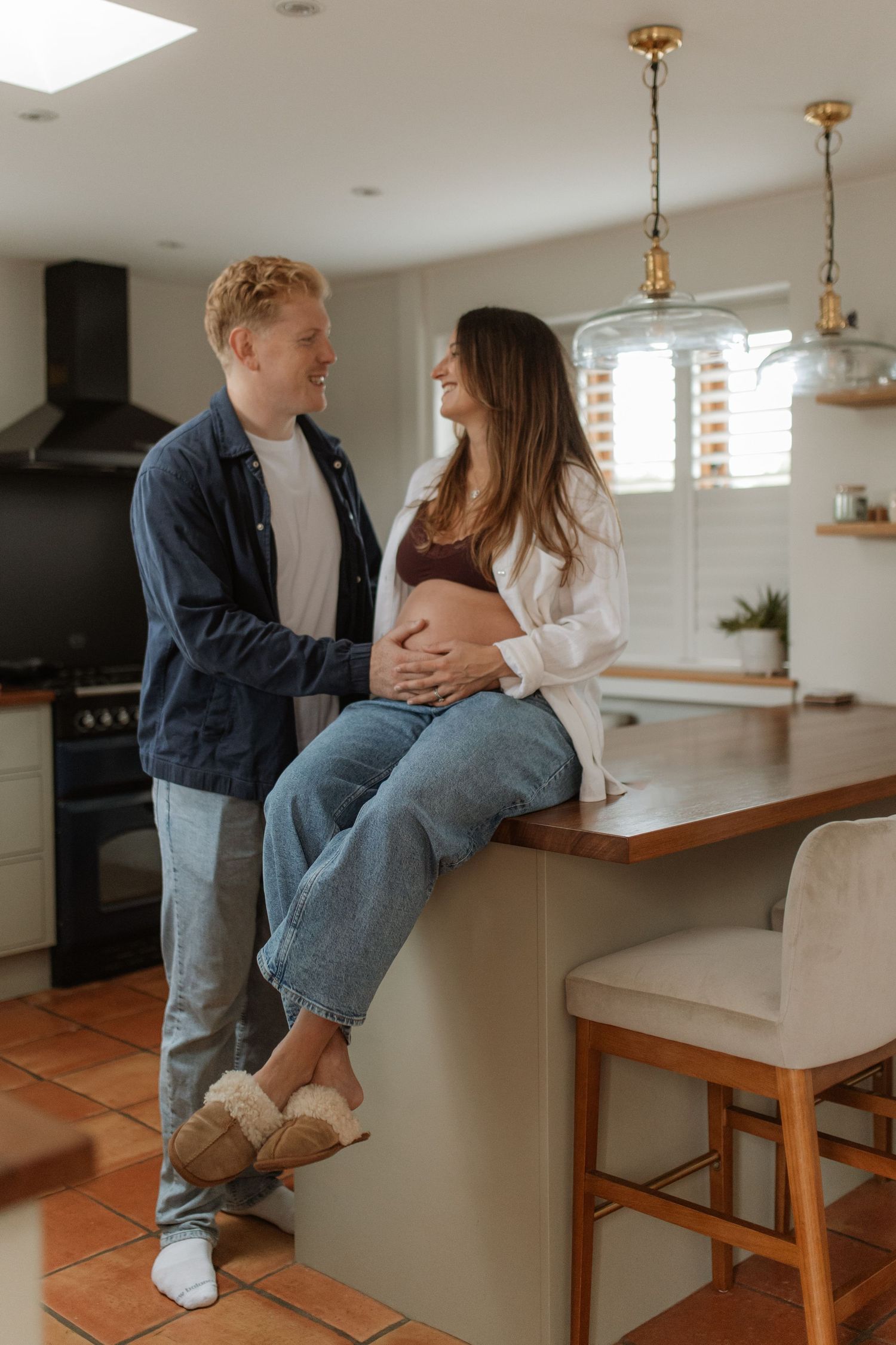 Happy couple in kitchen, man touching pregnant woman's baby bump while sitting on counter smiling.