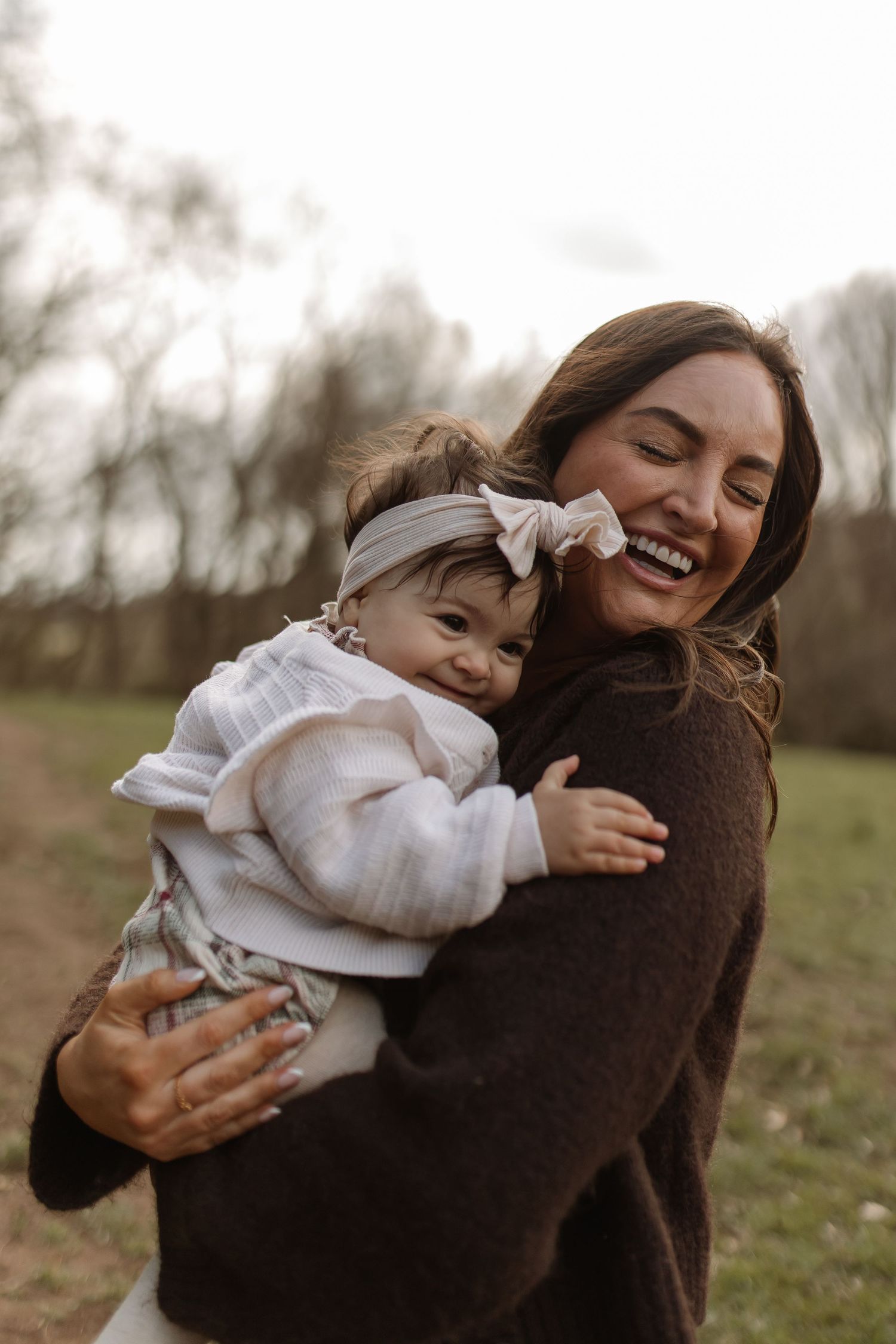 Joyful mother laughs while hugging her baby girl with a bow headband outdoors in a park.