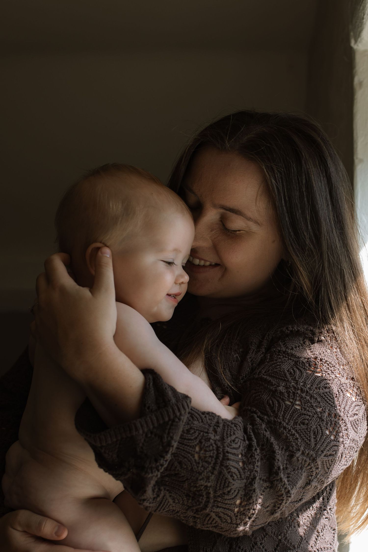 A smiling mother with long dark hair holding her happy baby close indoors.