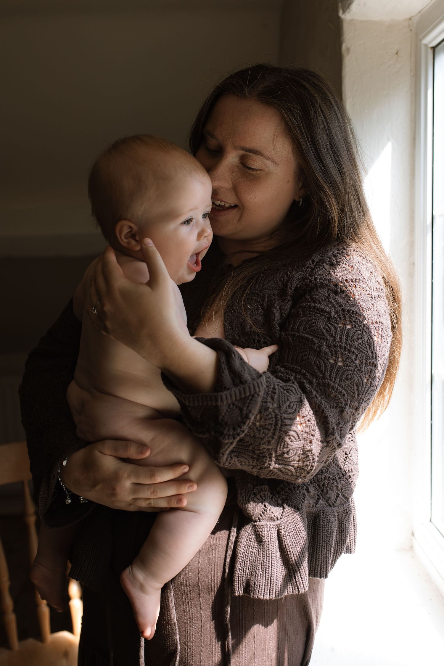 A joyful mother holds her laughing baby near a sunlit window indoors.