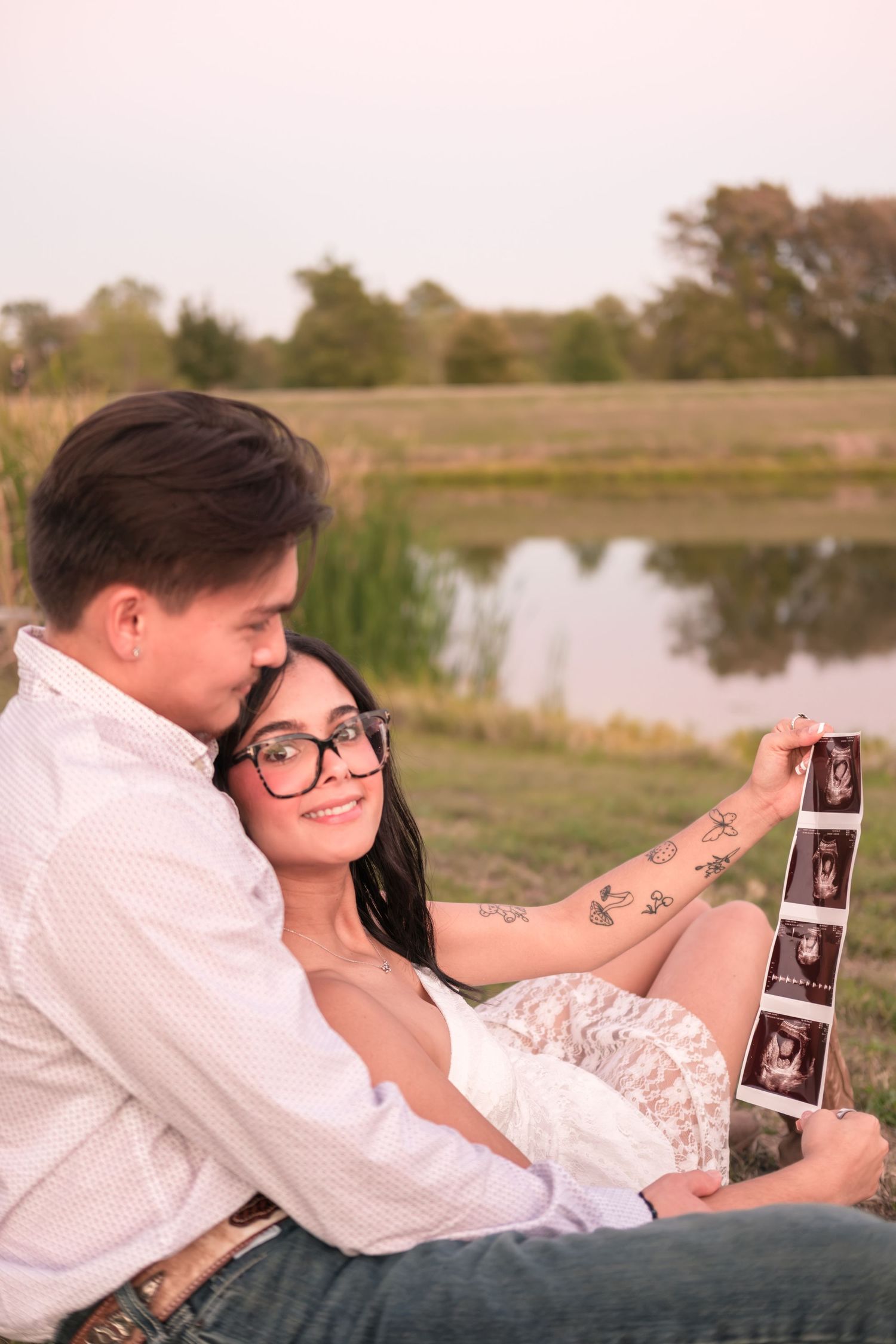 Couple sitting by a pond at sunset, woman with glasses smiling while holding ultrasound photos.