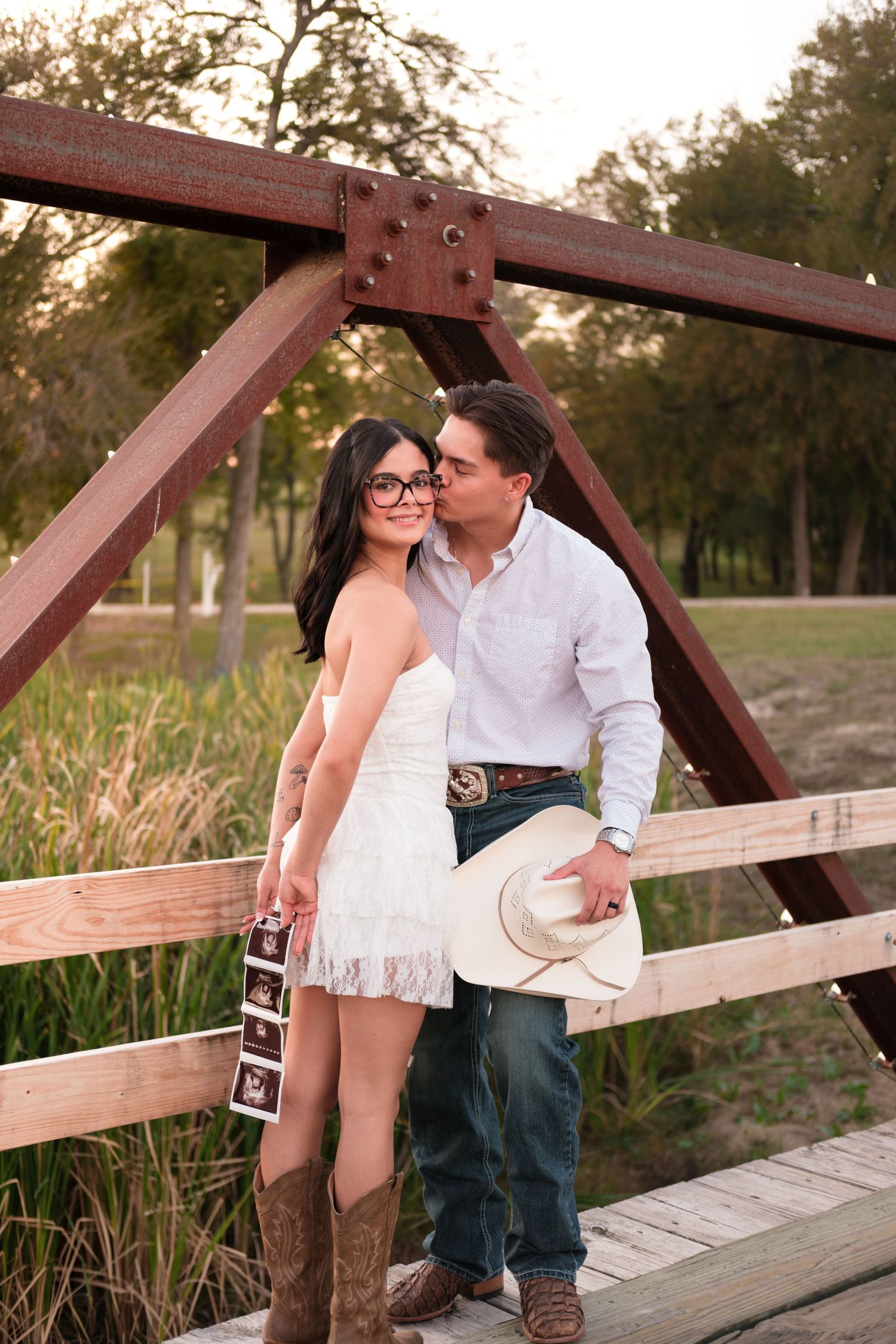 Couple on a wooden bridge at sunset, man kissing woman's cheek as she holds ultrasound photos.