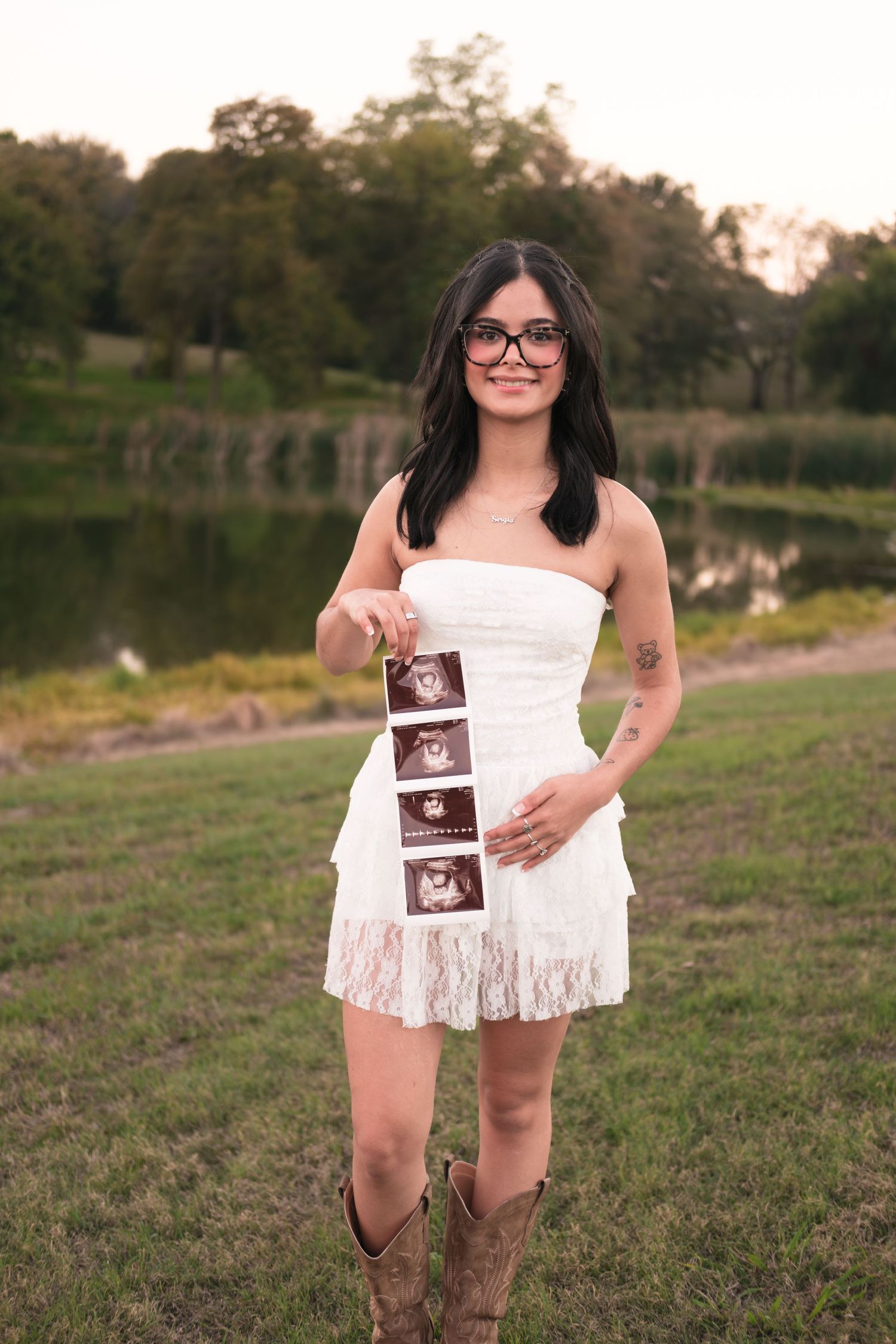 Smiling woman in white dress and cowboy boots holding ultrasound photos outdoors near a pond.