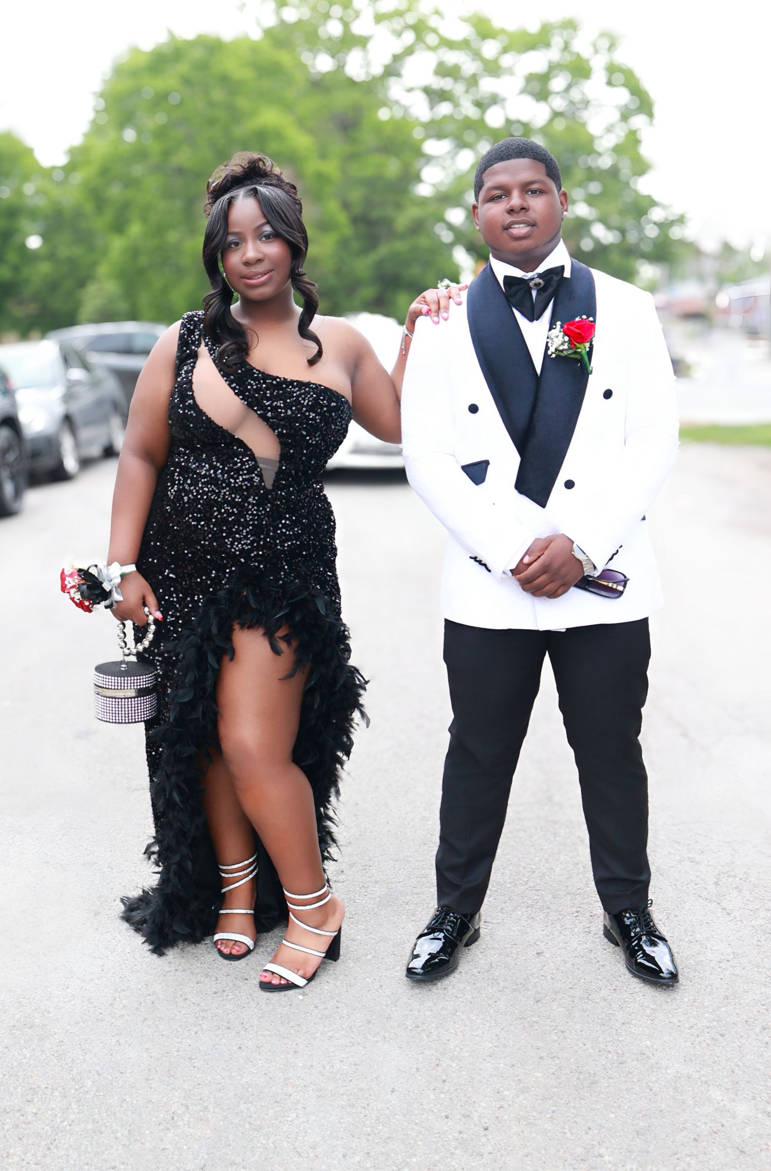 A couple dressed in formal prom attire poses outdoors, wearing black feathered gown and white tuxedo.