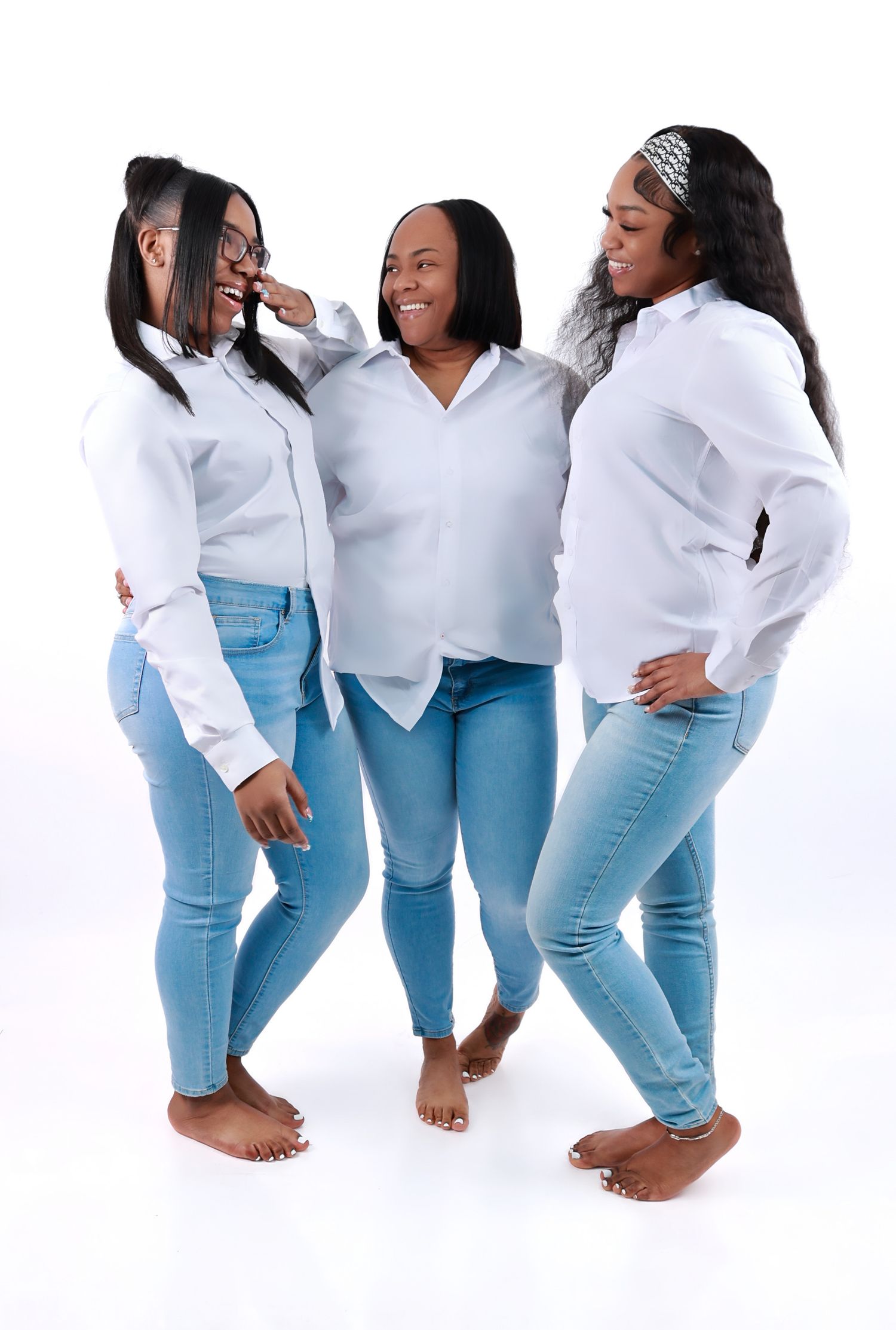Three women in matching white shirts and light blue jeans laugh together in a joyful group portrait.