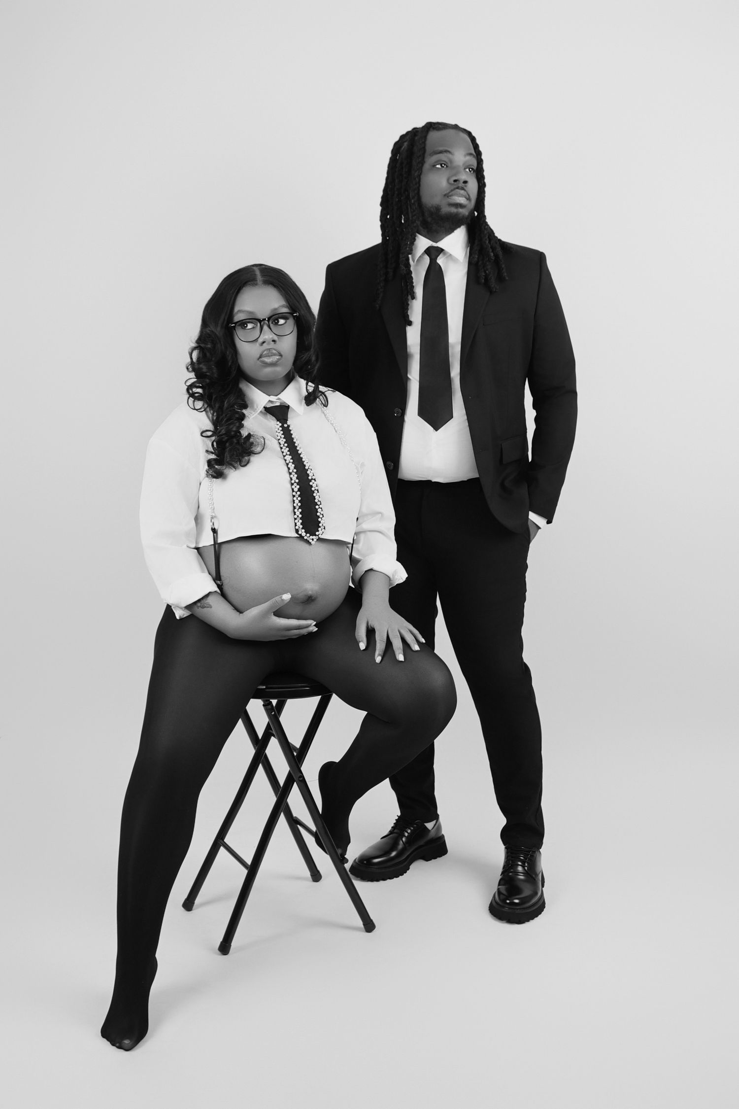 A black and white maternity portrait of a pregnant woman and suited man posing together in a professional studio setting.