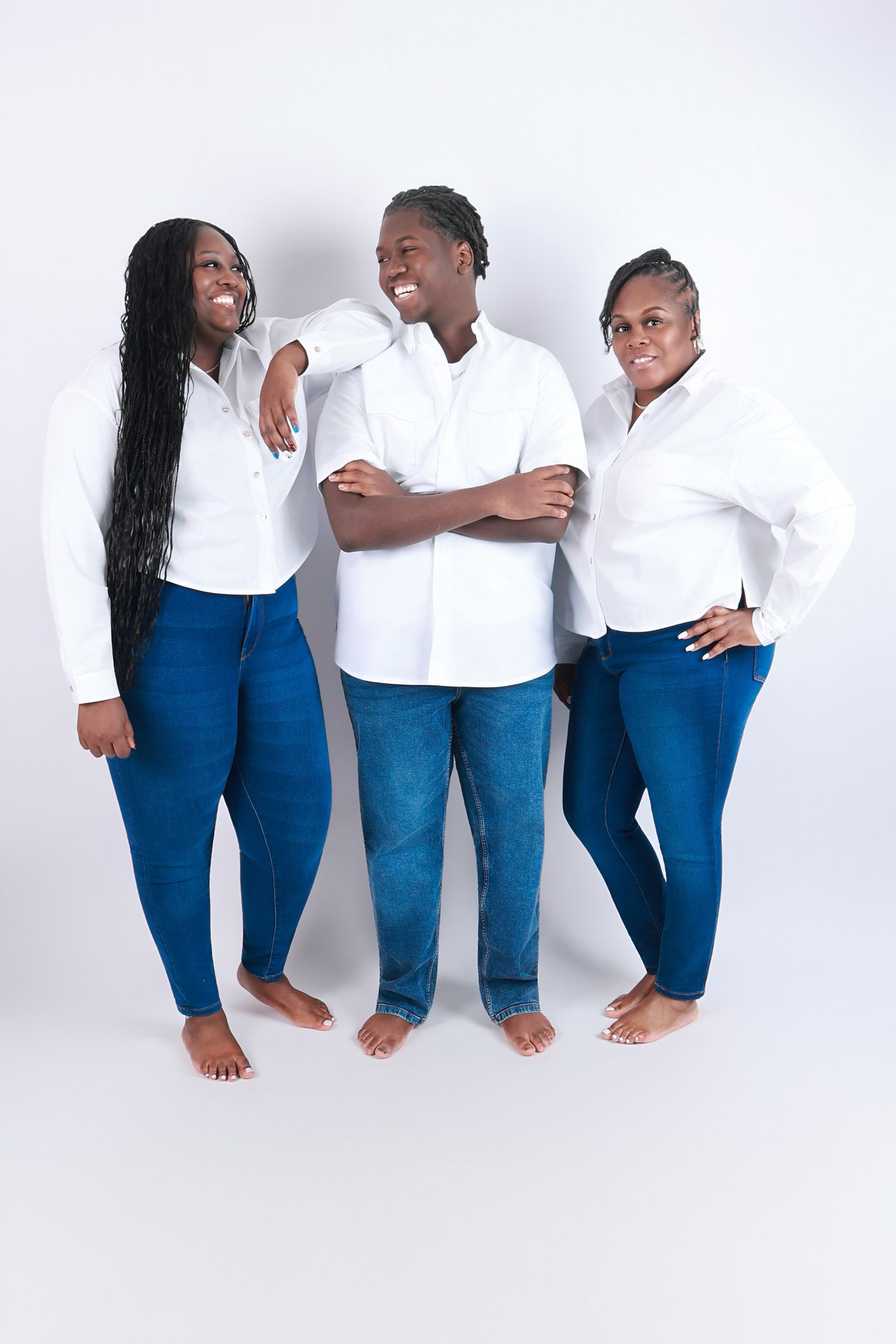 Three Black adults in matching white shirts and blue jeans pose barefoot in a white photo studio.