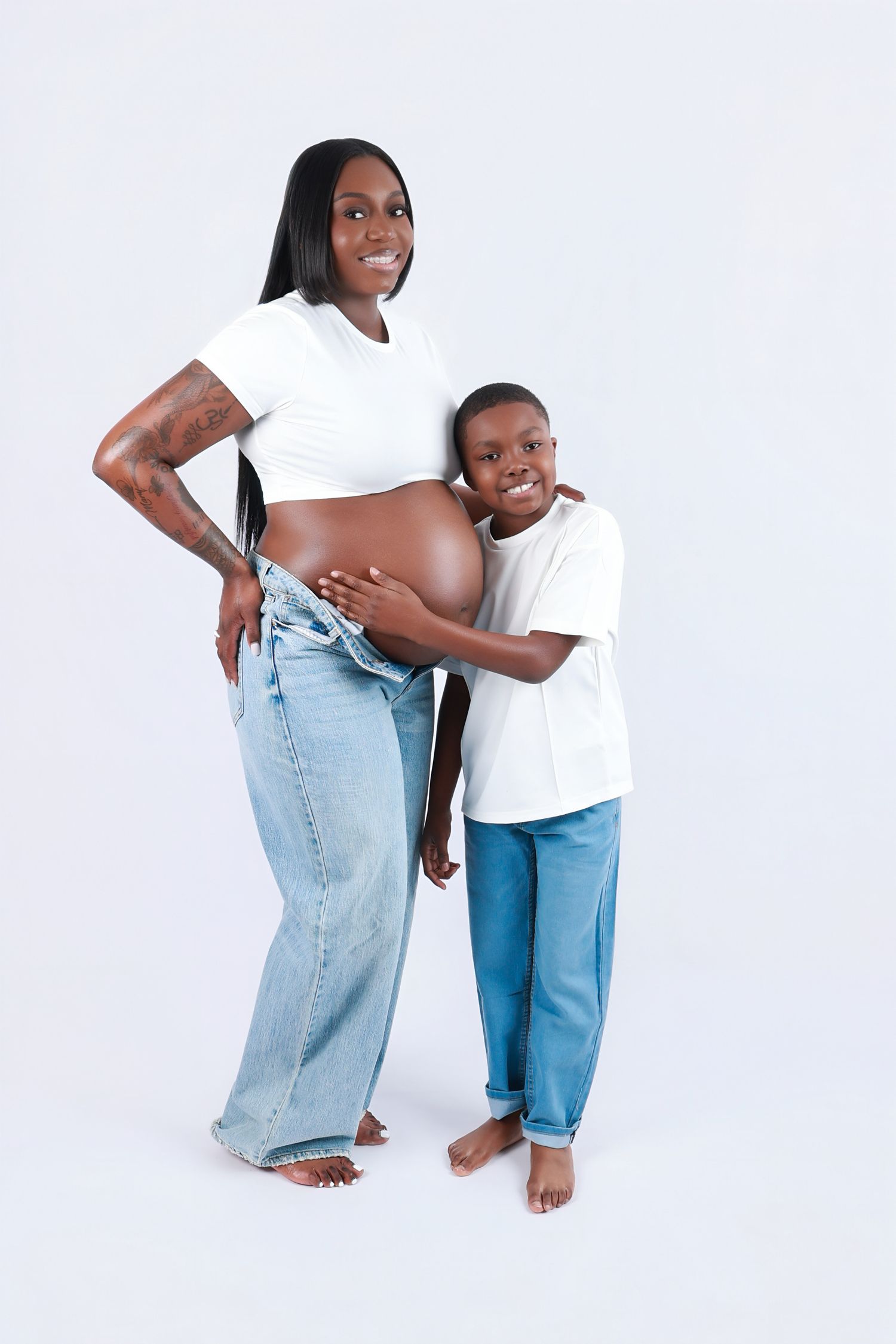 A pregnant Black woman in white crop top and jeans smiles as her young son hugs her baby bump.