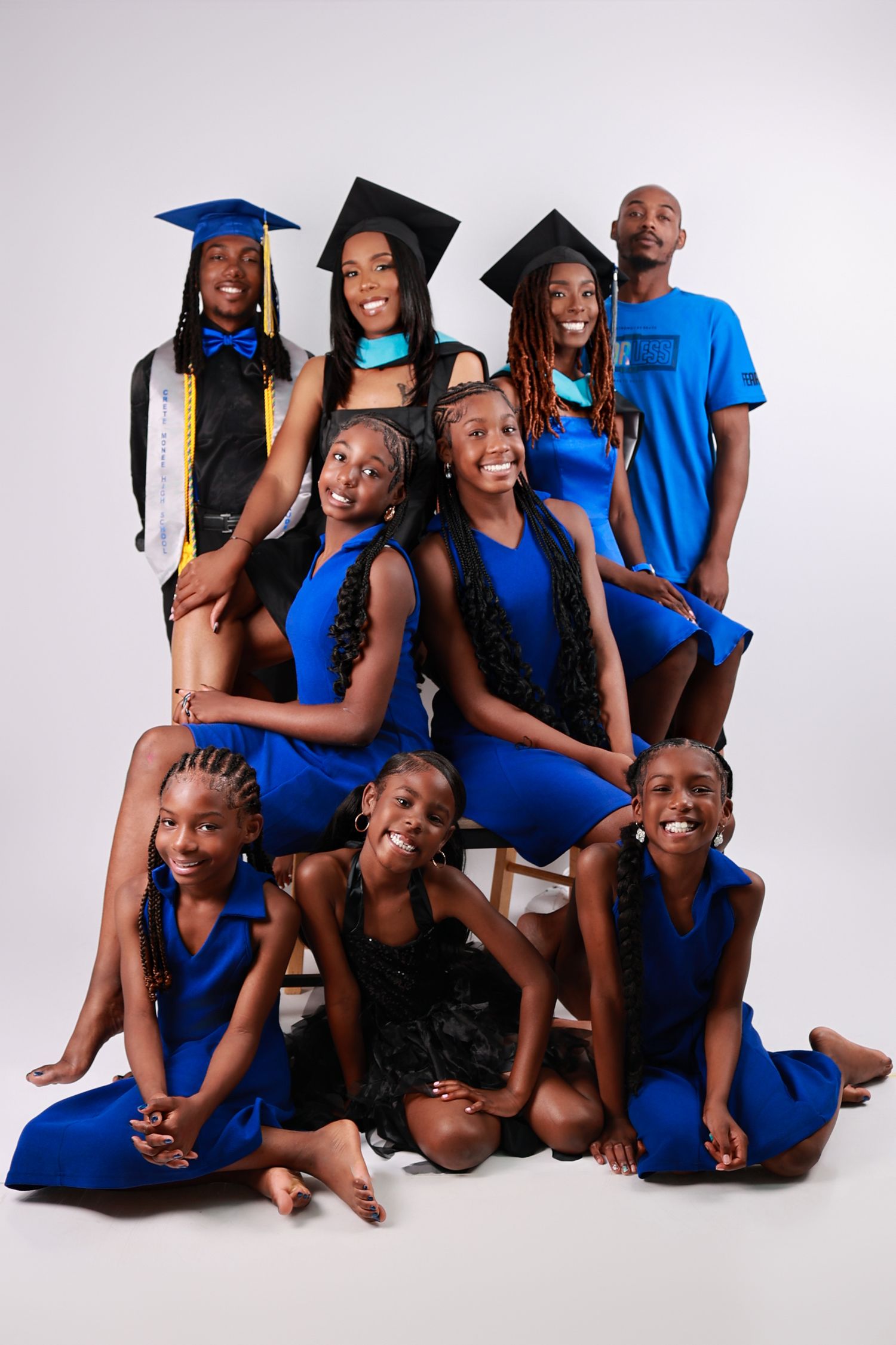 A large family group poses together in blue outfits with two members wearing graduation caps and gowns in a studio portrait.