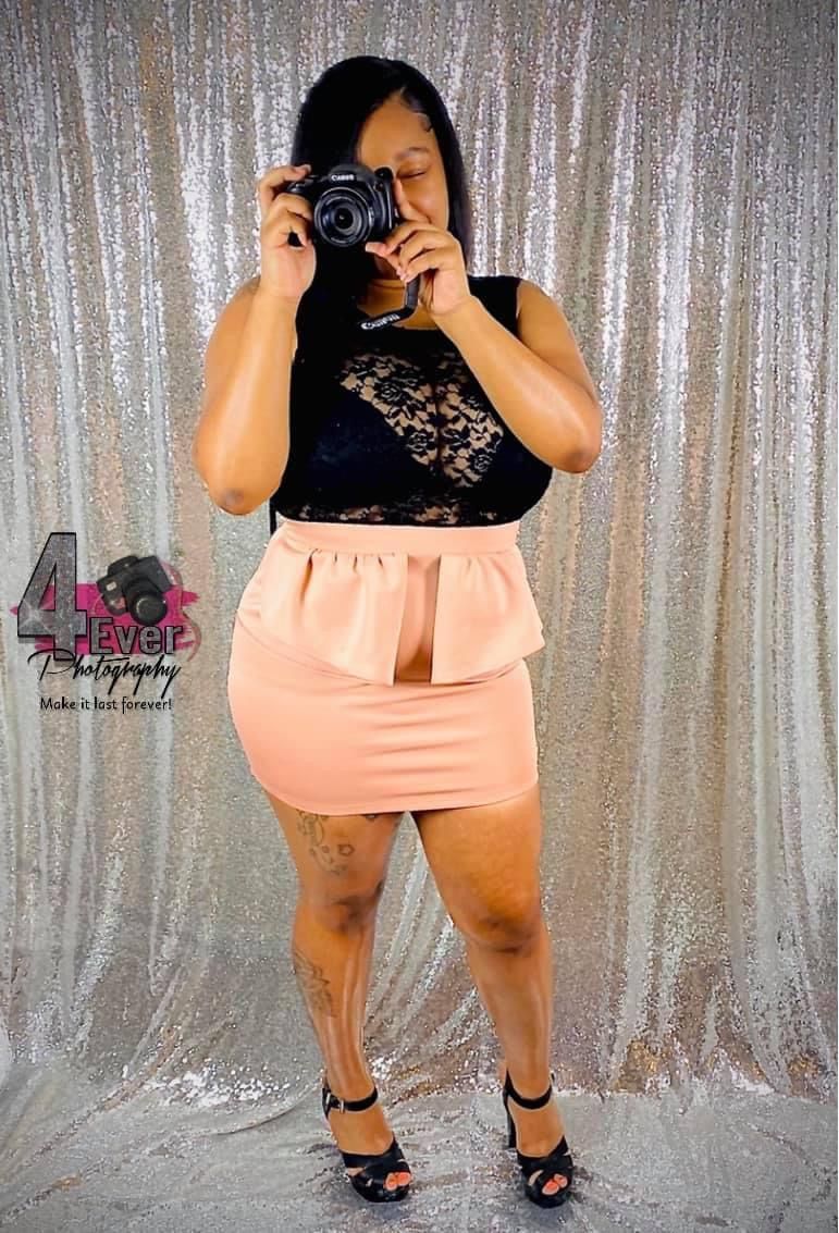Woman in black lace top and peach peplum skirt taking mirror selfie with DSLR camera.