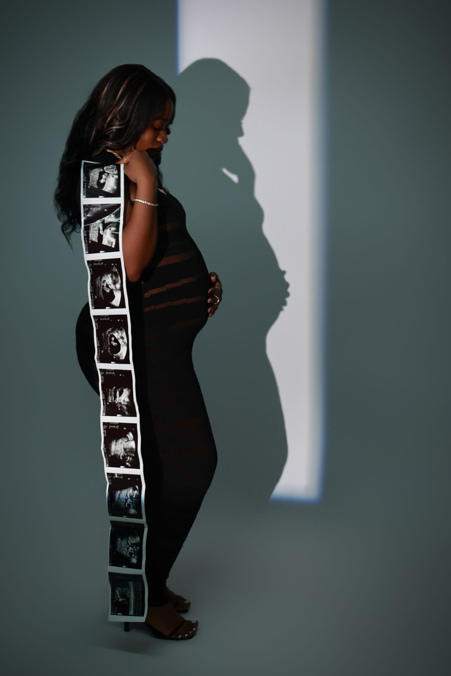 Pregnant woman in black dress holding ultrasound strip beside dramatic shadow on gray wall.