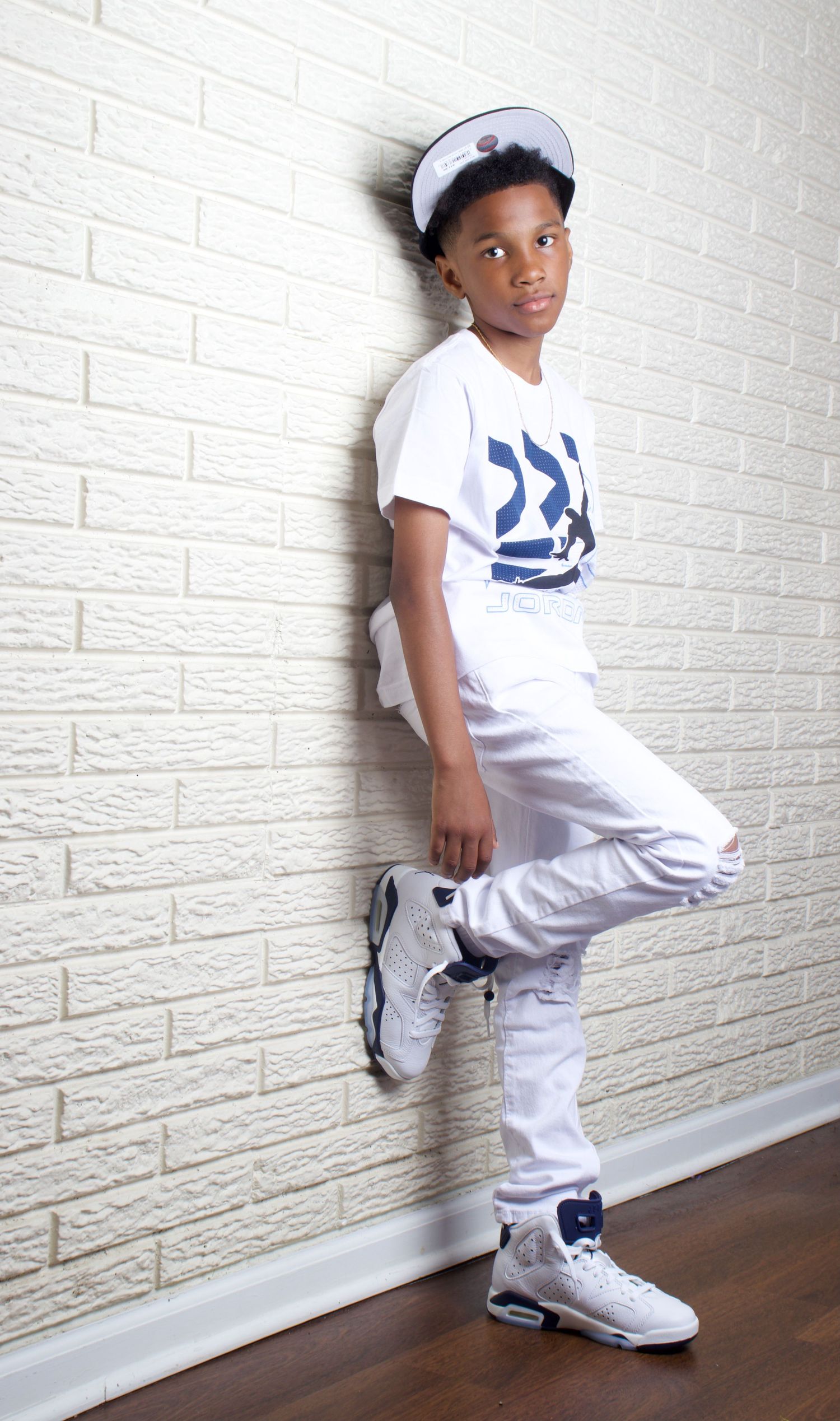 Stylish young boy in white outfit and cap leaning against white brick wall.