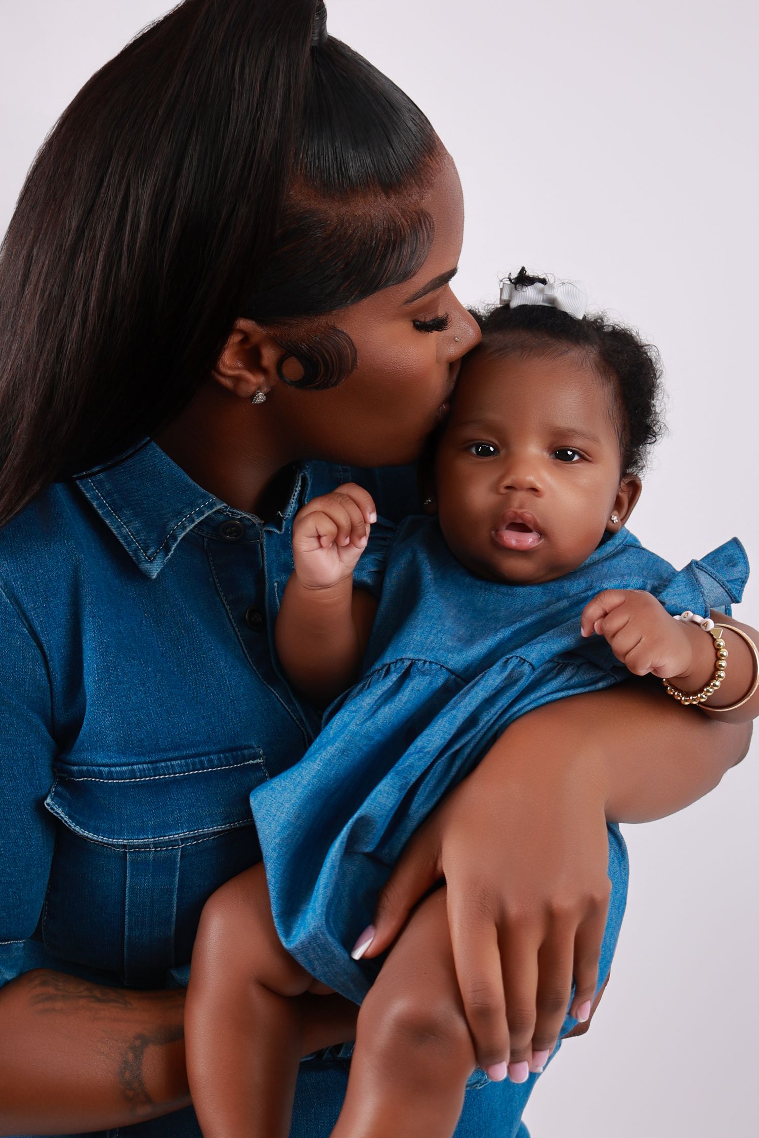 Mother in denim outfit tenderly kissing her baby girl dressed in matching denim dress.
