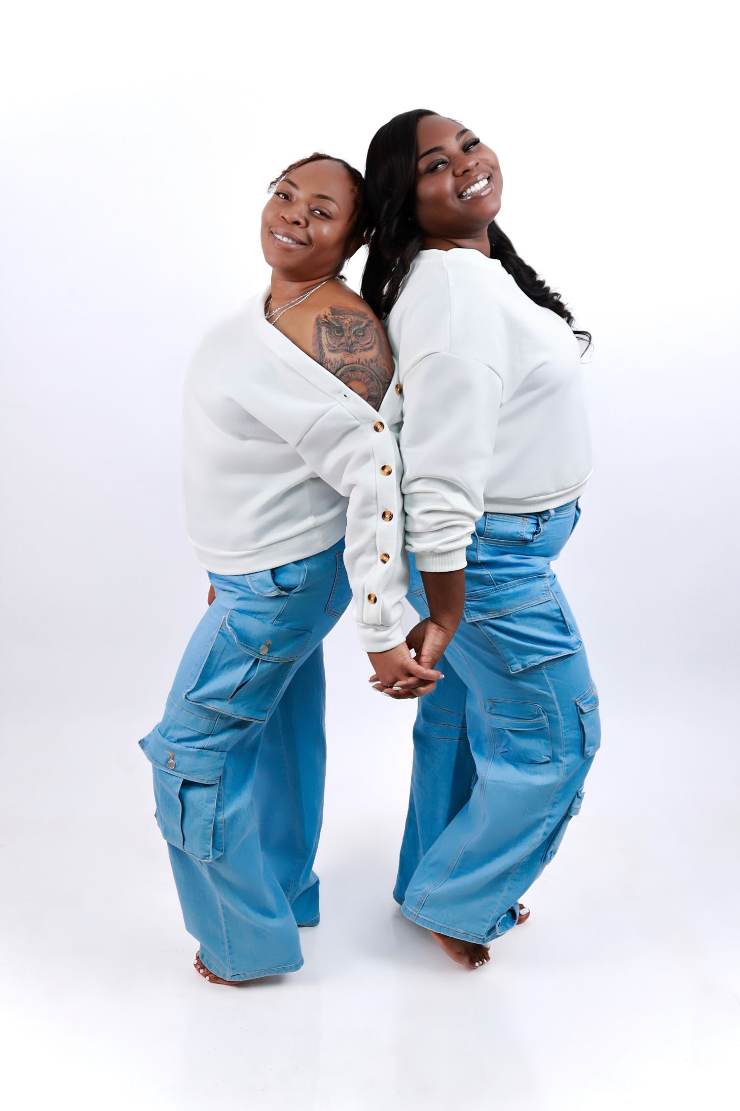 Two women in matching white tops and blue cargo jeans pose back-to-back in a studio photo shoot.