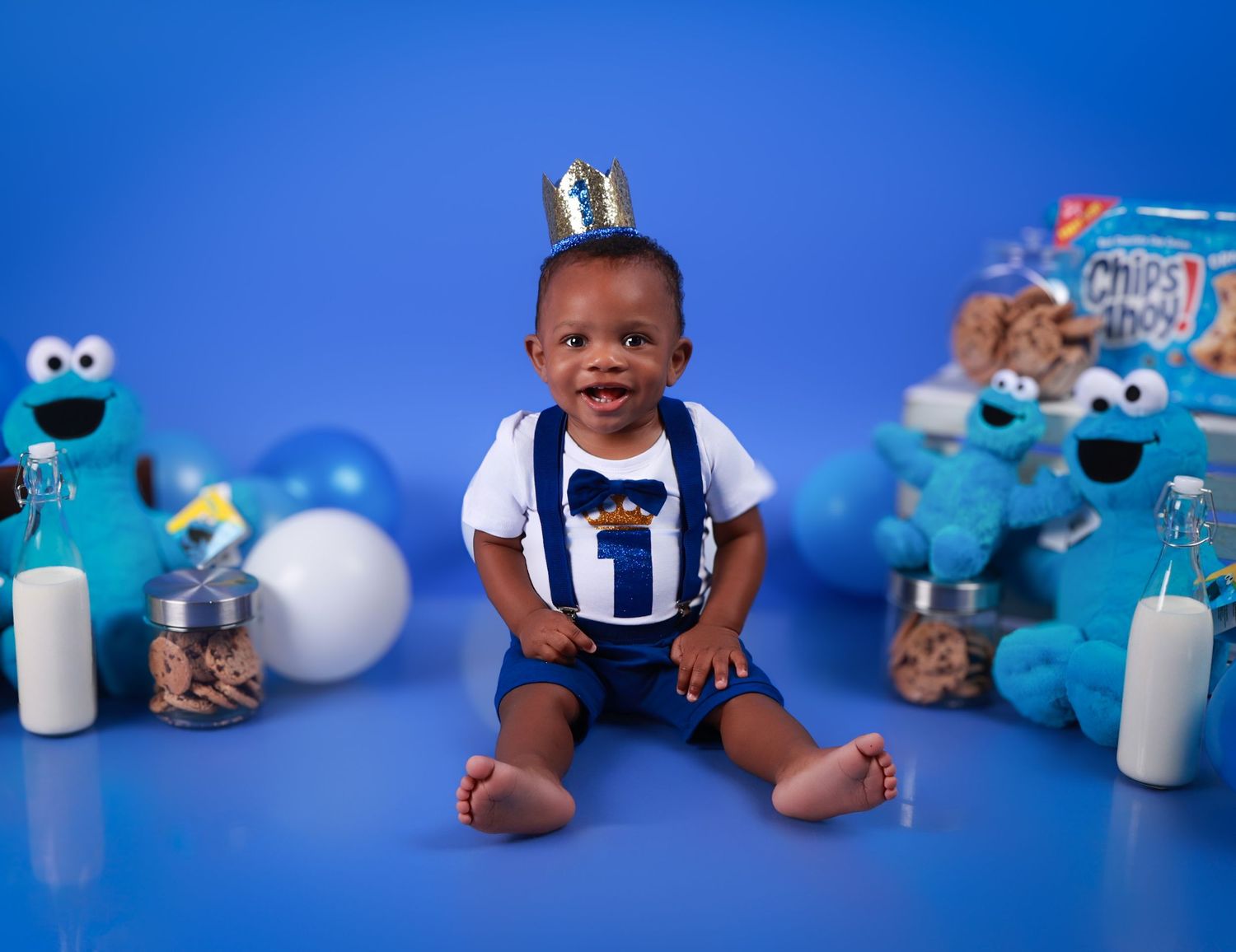 Baby boy in blue and white outfit with gold crown sits surrounded by Cookie Monster toys for a first birthday cake smash.