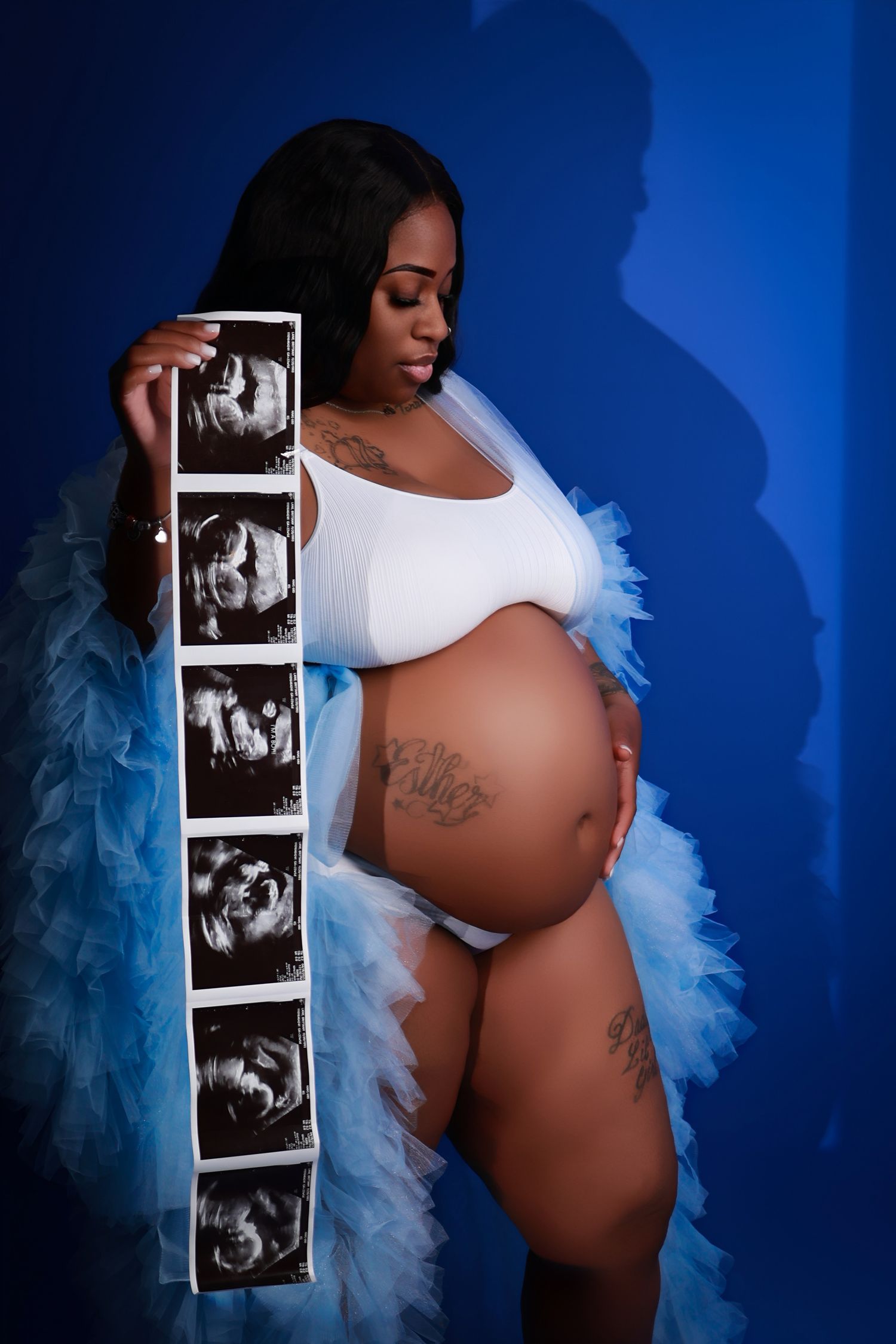 Pregnant woman in white bodysuit holding ultrasound photos wearing a blue feathered robe.