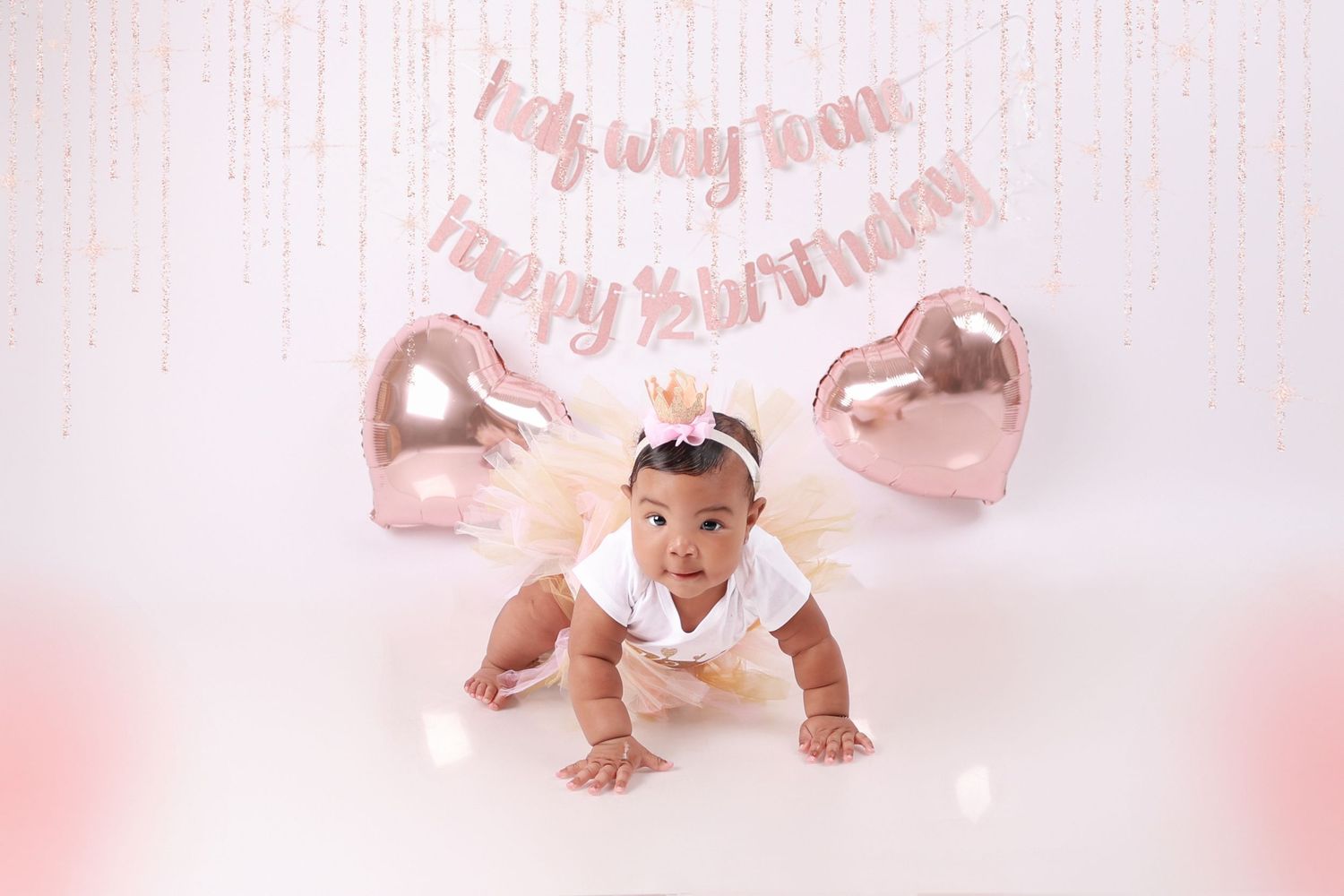 Baby girl in white outfit crawls under half birthday banner and rose gold heart balloons in a soft pink studio setting.