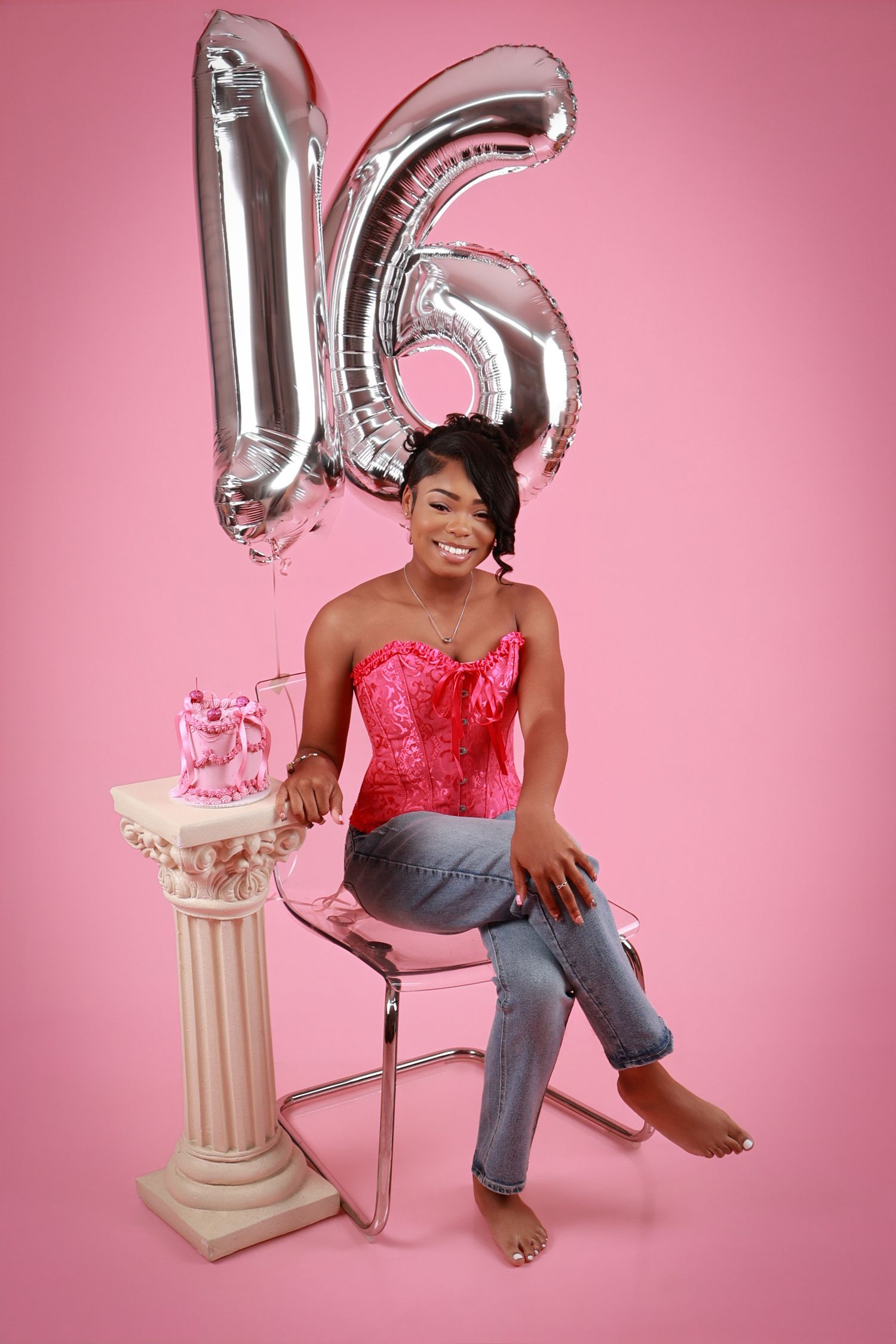 Teen girl celebrating sweet 16 birthday with silver balloon numbers and pink cake on pink background.