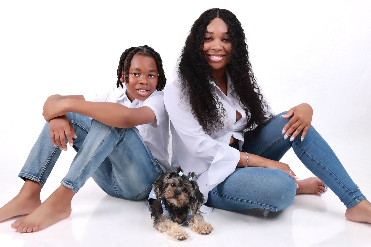 A woman and boy sit together on white floor with small dog, both wearing white tops and jeans.