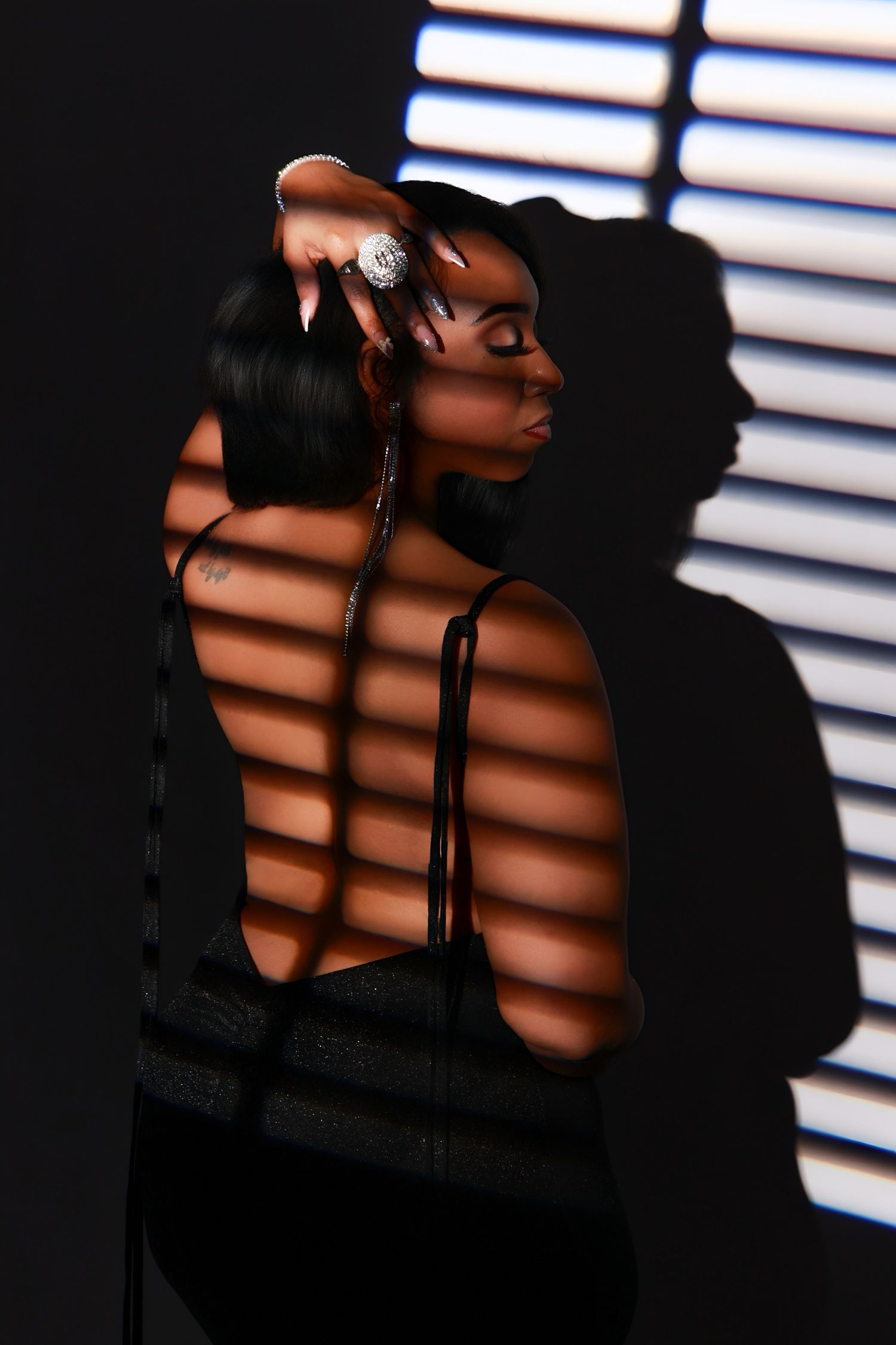 Woman posing with dramatic shadow stripes from blinds on dark background wearing black backless dress.