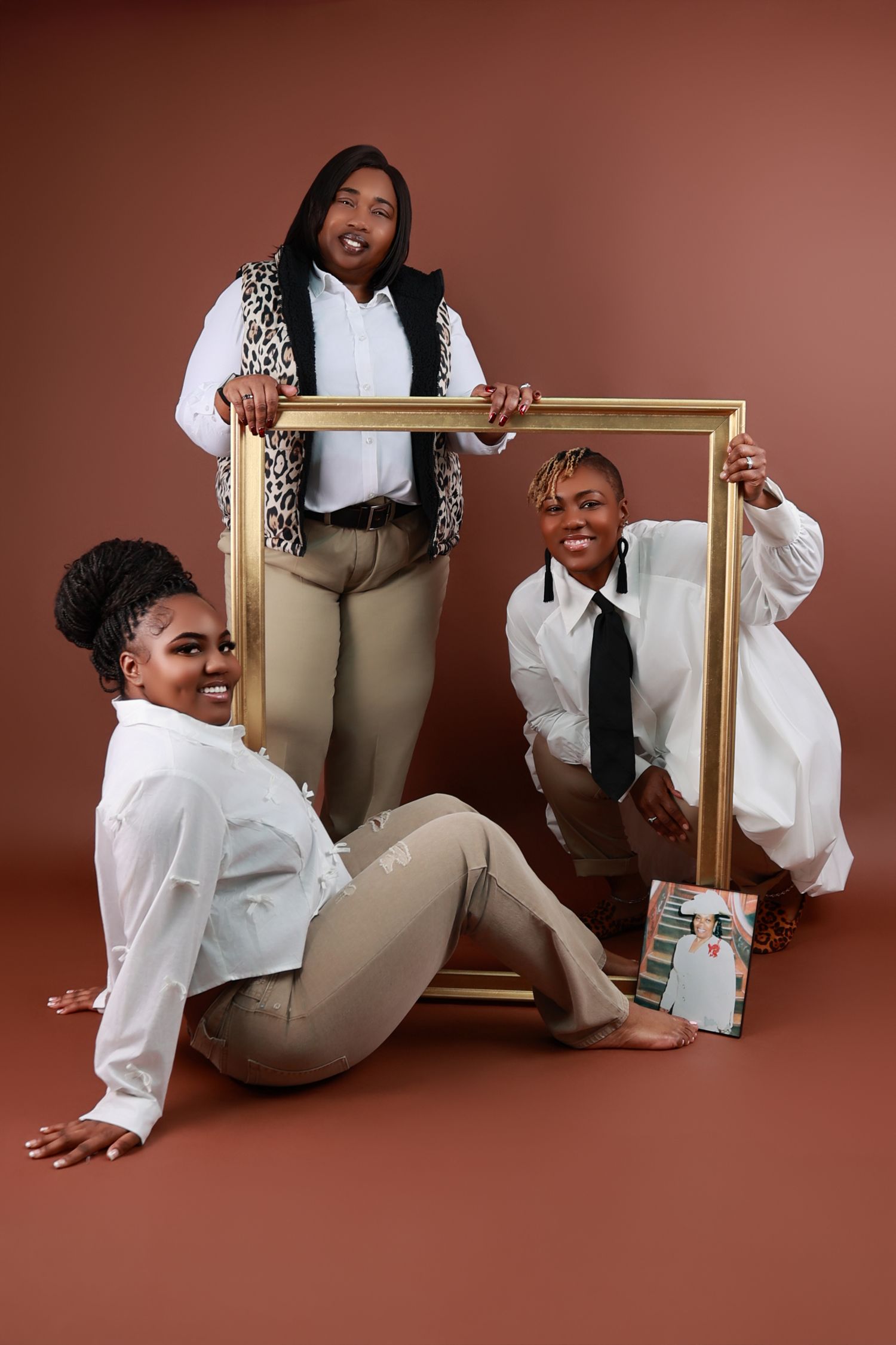 Three women pose with a gold picture frame holding a framed photo in a warm brown studio setting.
