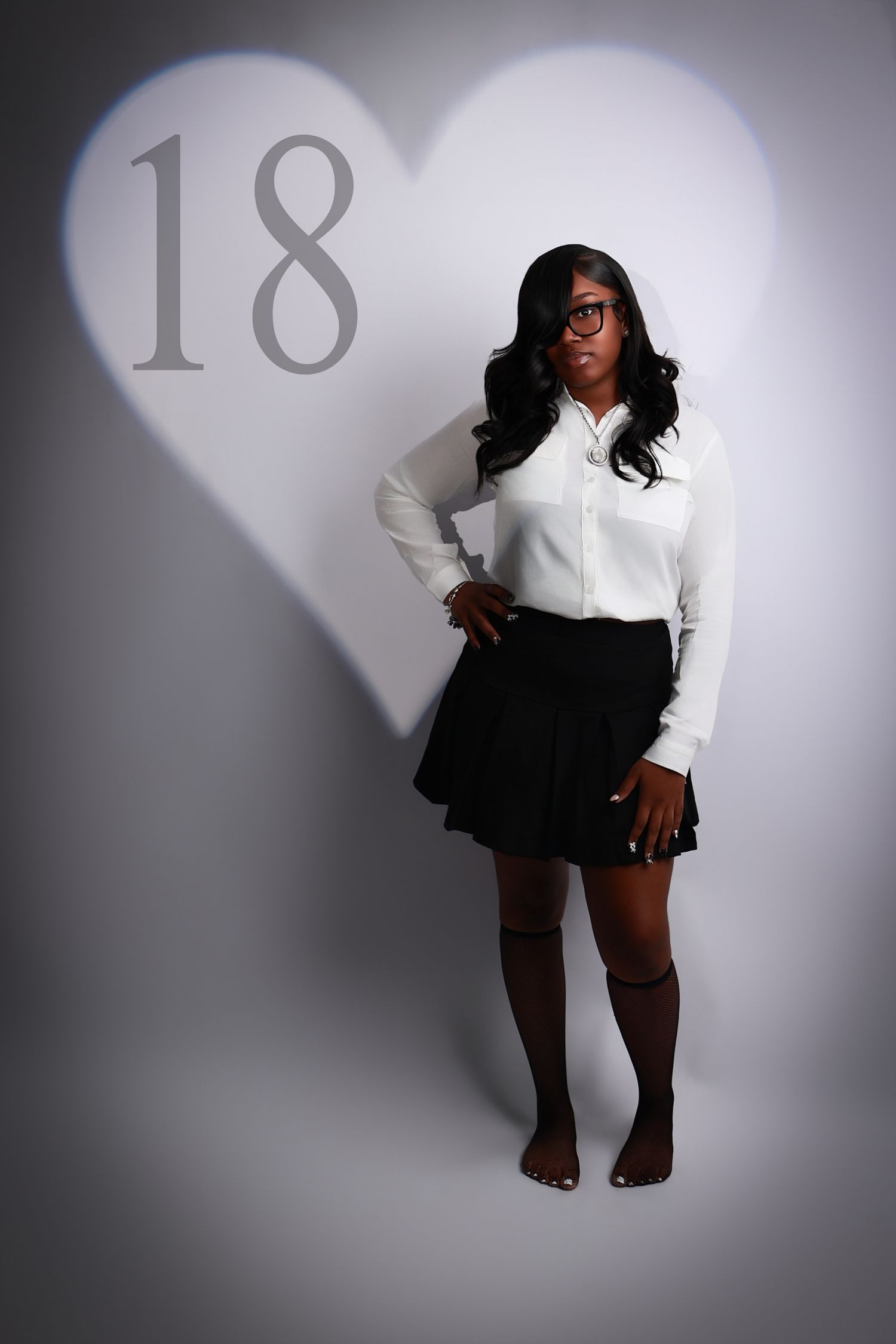 Young woman in white shirt and black skirt poses in front of a heart-shaped spotlight with the number 18 for her birthday.