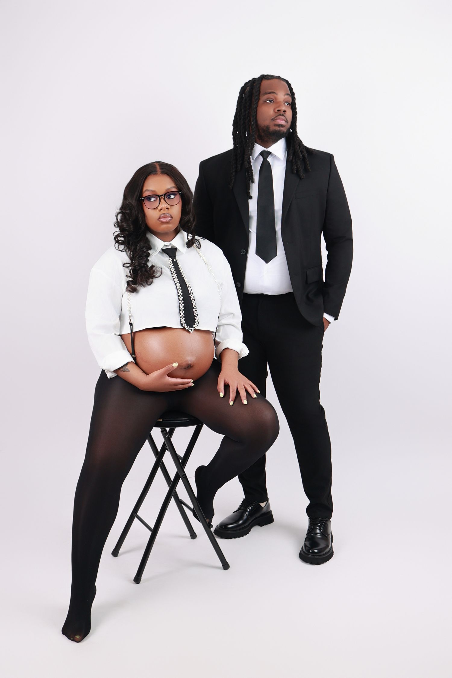 A pregnant Black woman and her partner pose formally in business attire for a maternity photo shoot.