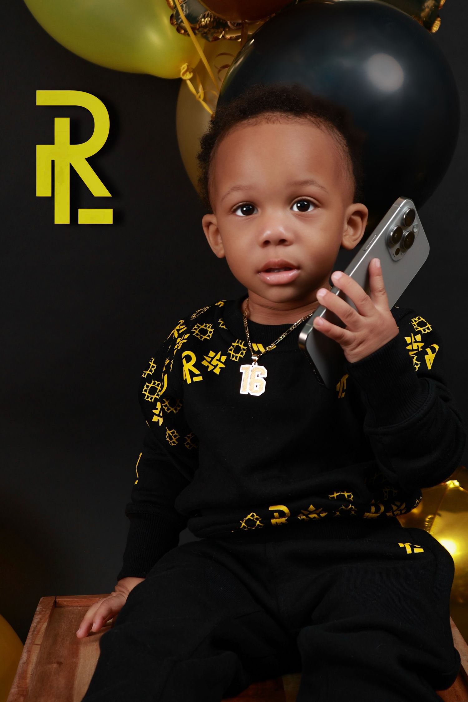 Toddler boy in black and gold outfit holding phone, seated with balloons on dark background.