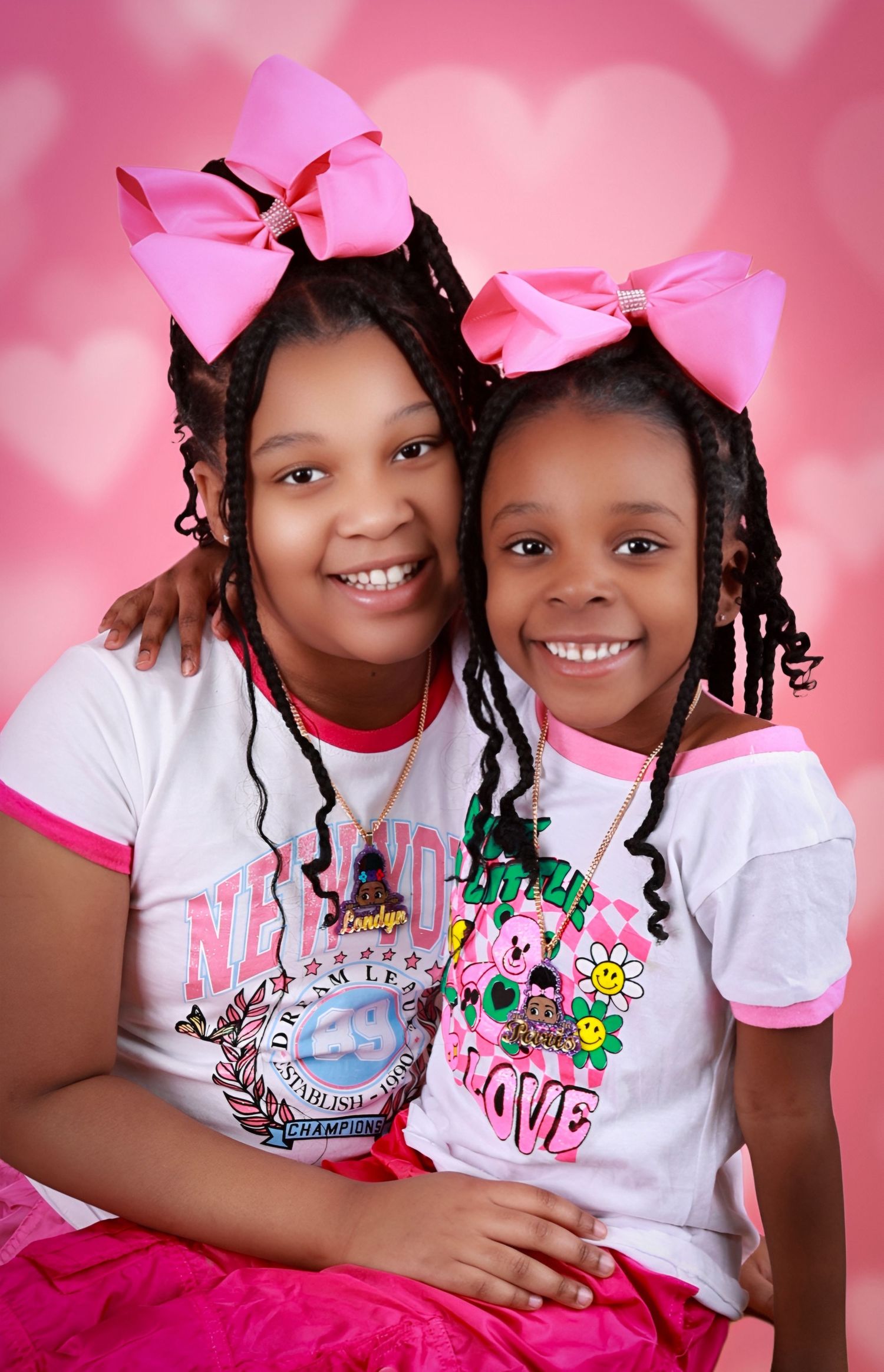 Two young girls in matching pink bows and outfits smile together against a pink heart background.