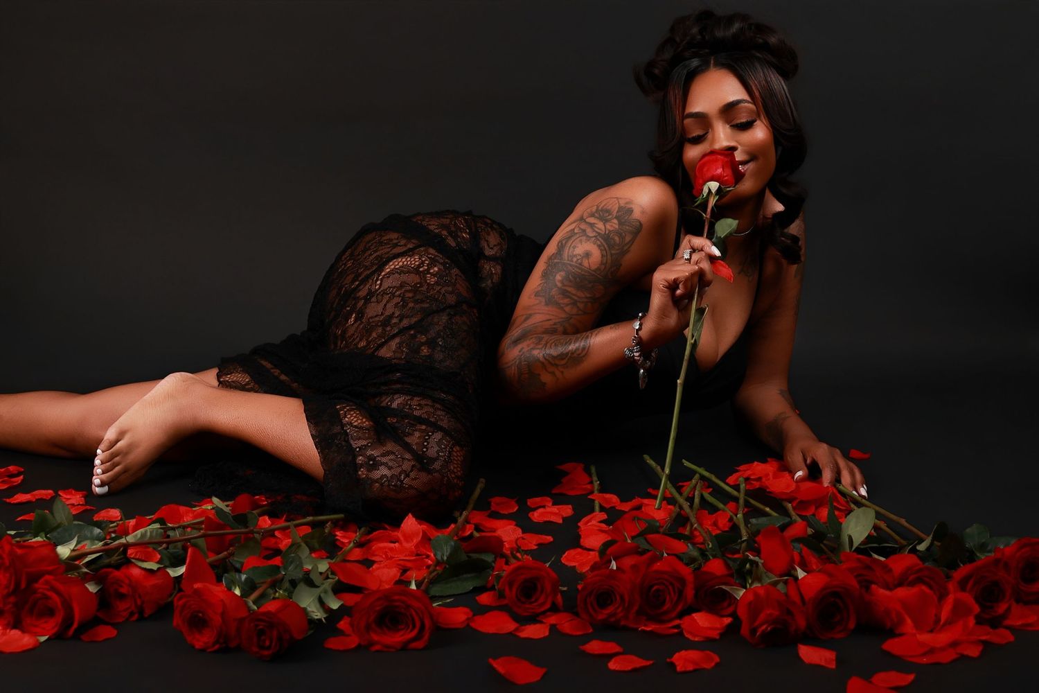Woman in black lace dress lying among red roses in a dark studio setting.