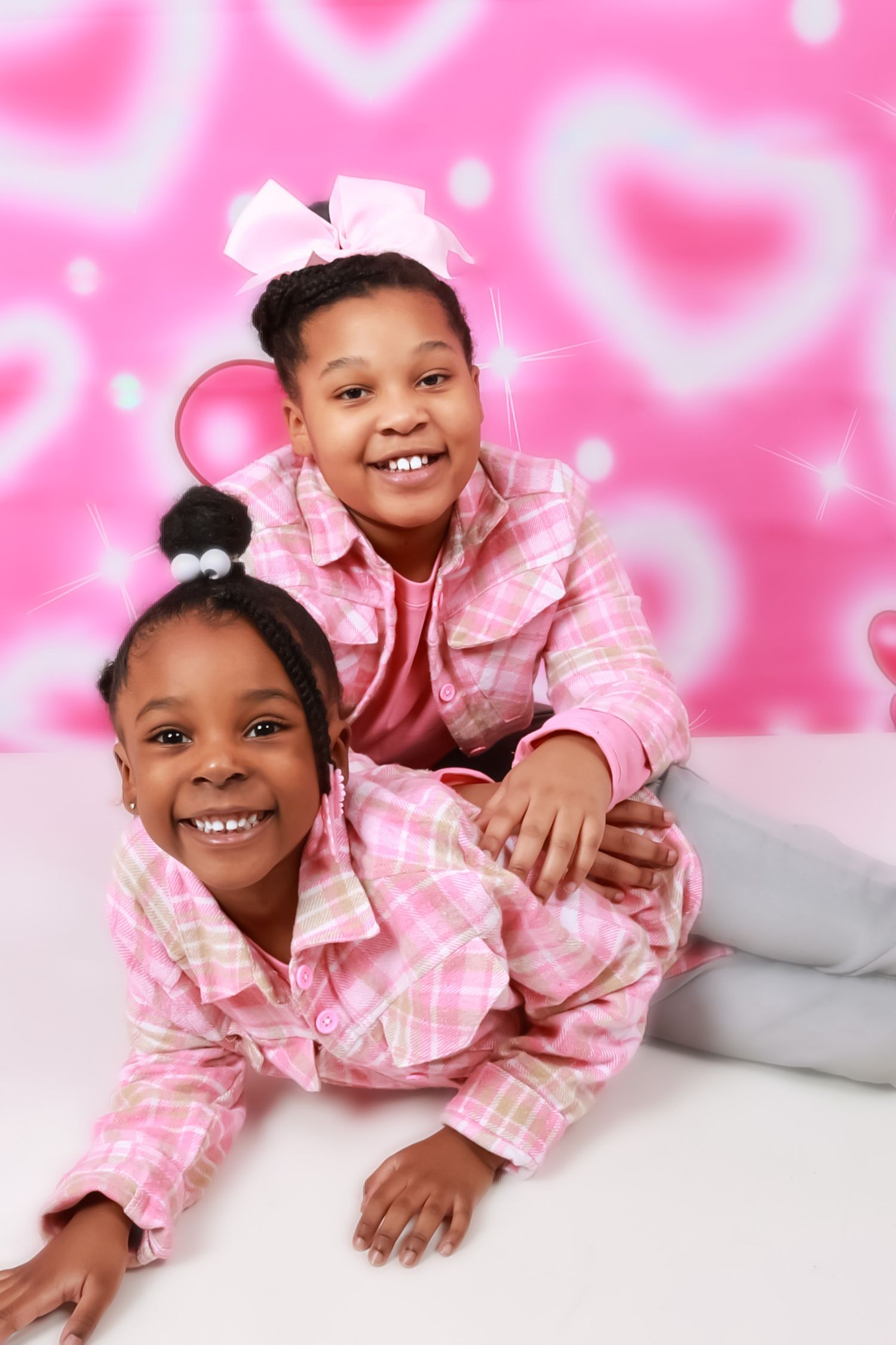 Two young sisters smiling in matching pink plaid outfits against pink heart background.