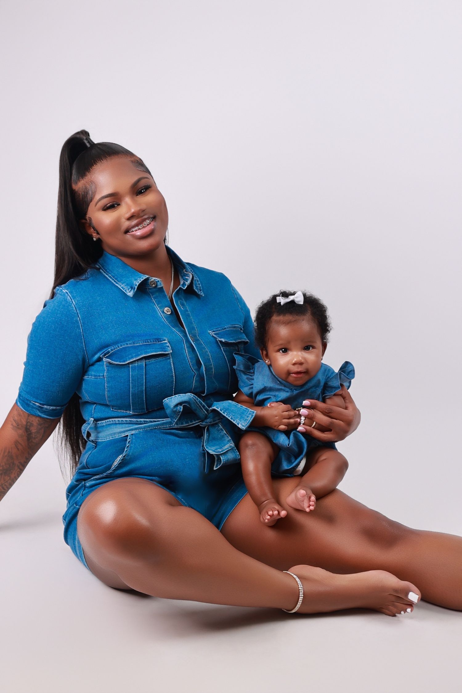 Smiling mother in denim romper sitting with infant daughter in matching denim outfit.