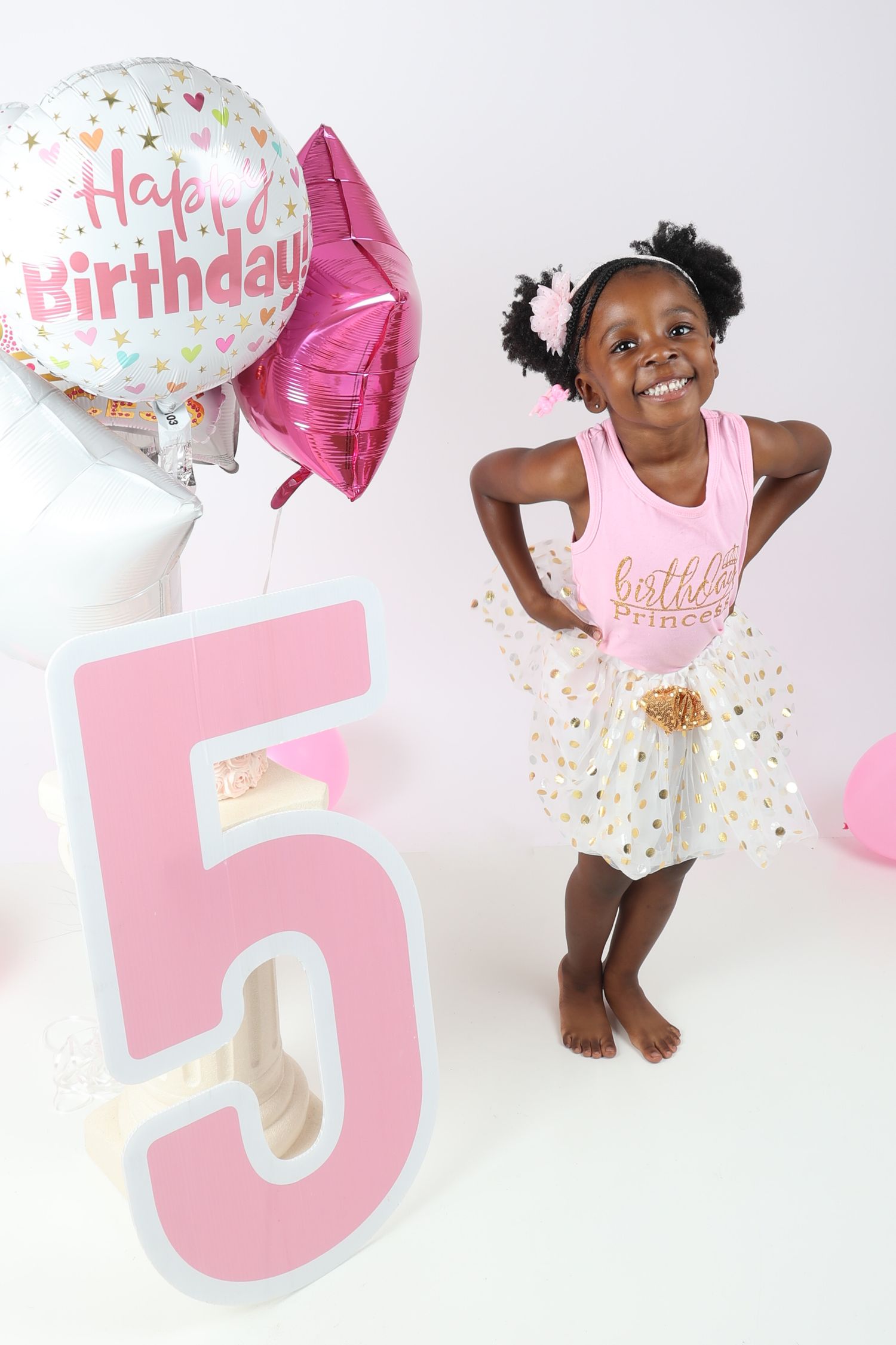 Happy girl celebrating 5th birthday with pink tutu, balloons, and large number 5 decoration.