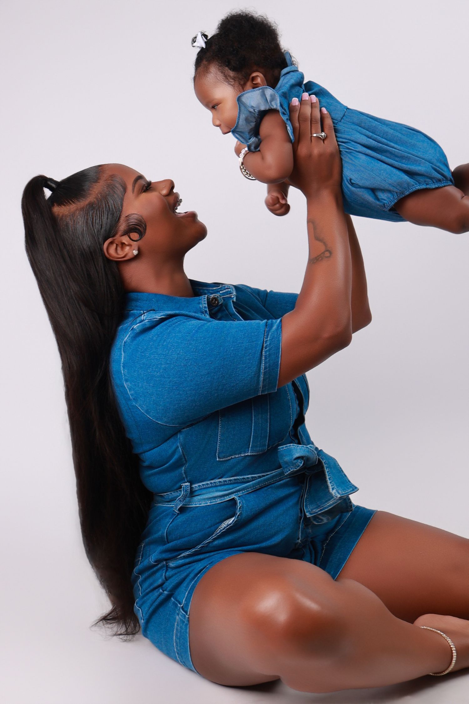 A woman in a denim dress lifts her smiling baby girl overhead in a joyful studio photo.