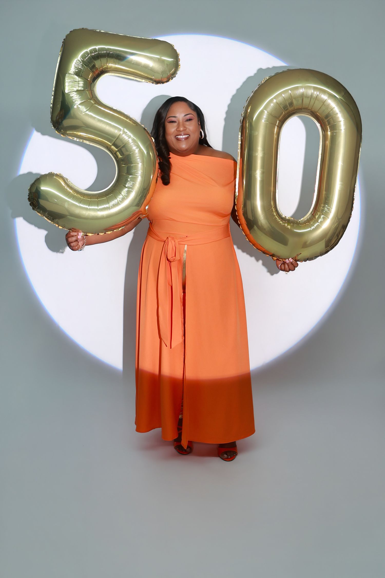 A woman in an orange dress celebrates her 50th birthday holding gold number balloon digits.
