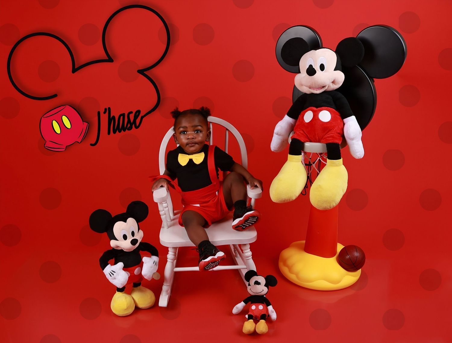 Toddler in Mickey Mouse outfit sitting in white chair surrounded by Mickey Mouse plush toys.