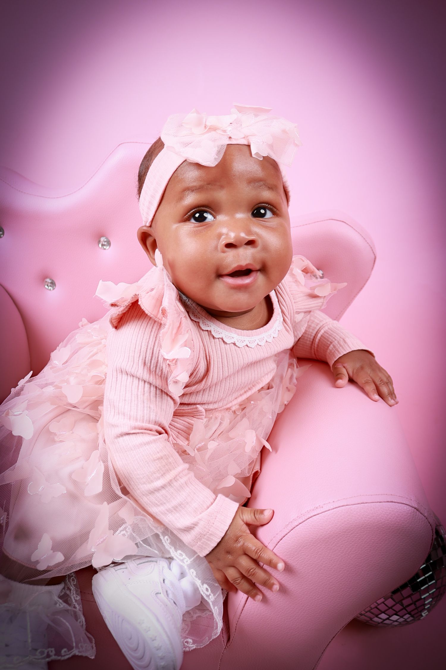 Adorable baby girl in pink dress and bow headband sitting in pink velvet chair.