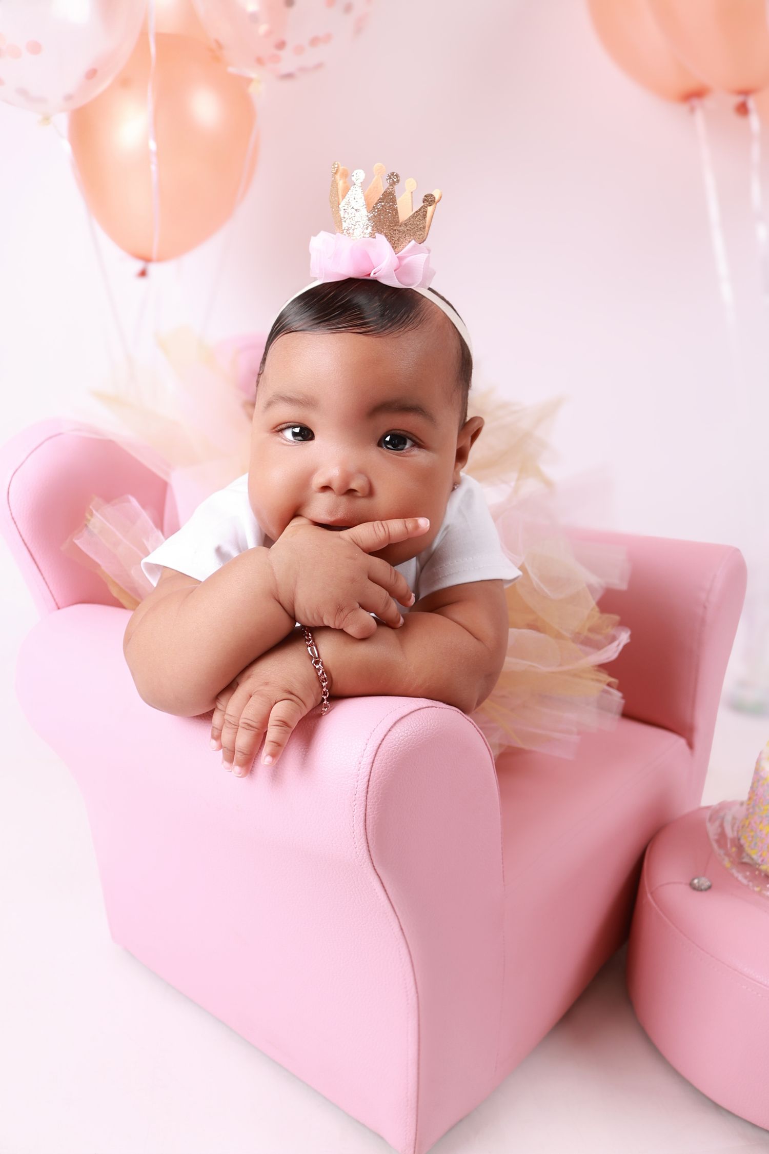 Baby girl in pink tutu and gold crown sitting in a pink chair surrounded by rose gold balloons.