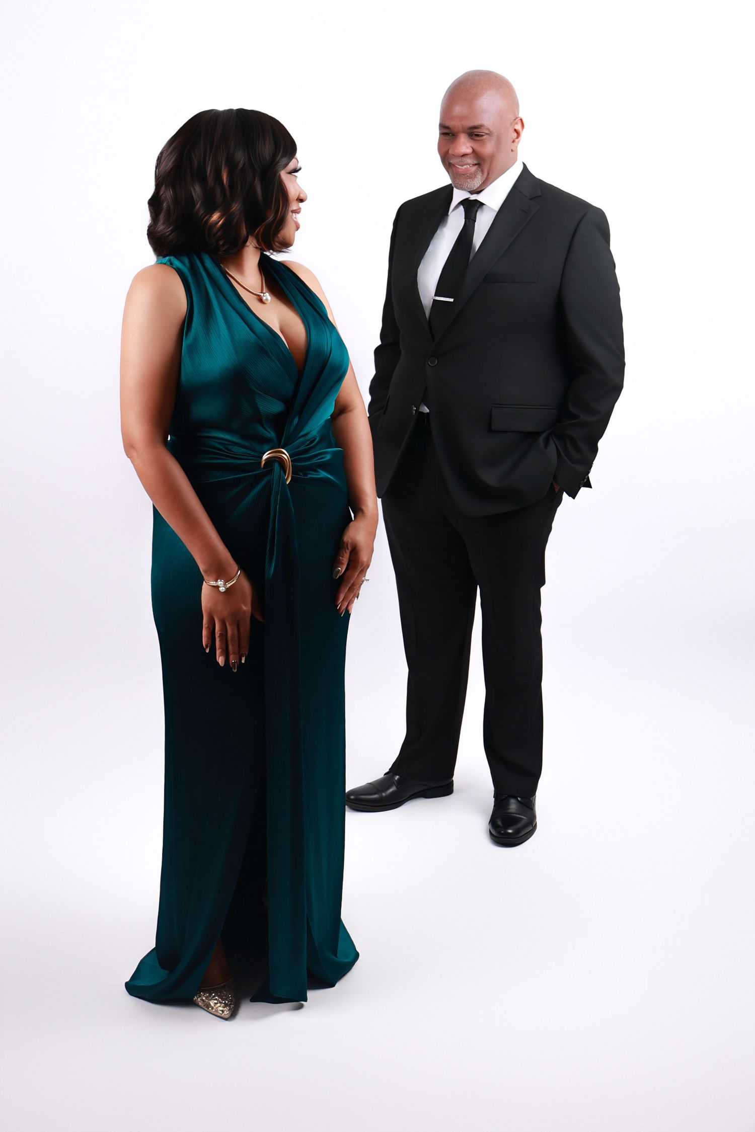 An elegant couple poses together, woman in teal gown and man in black suit, on white background.