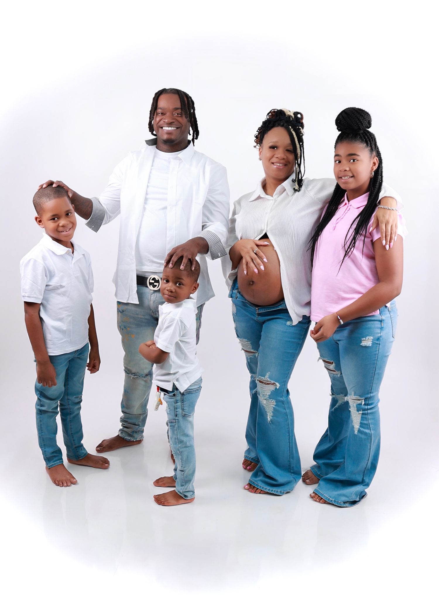 A family of five in white shirts and jeans poses together, with a pregnant mother showing her baby bump.