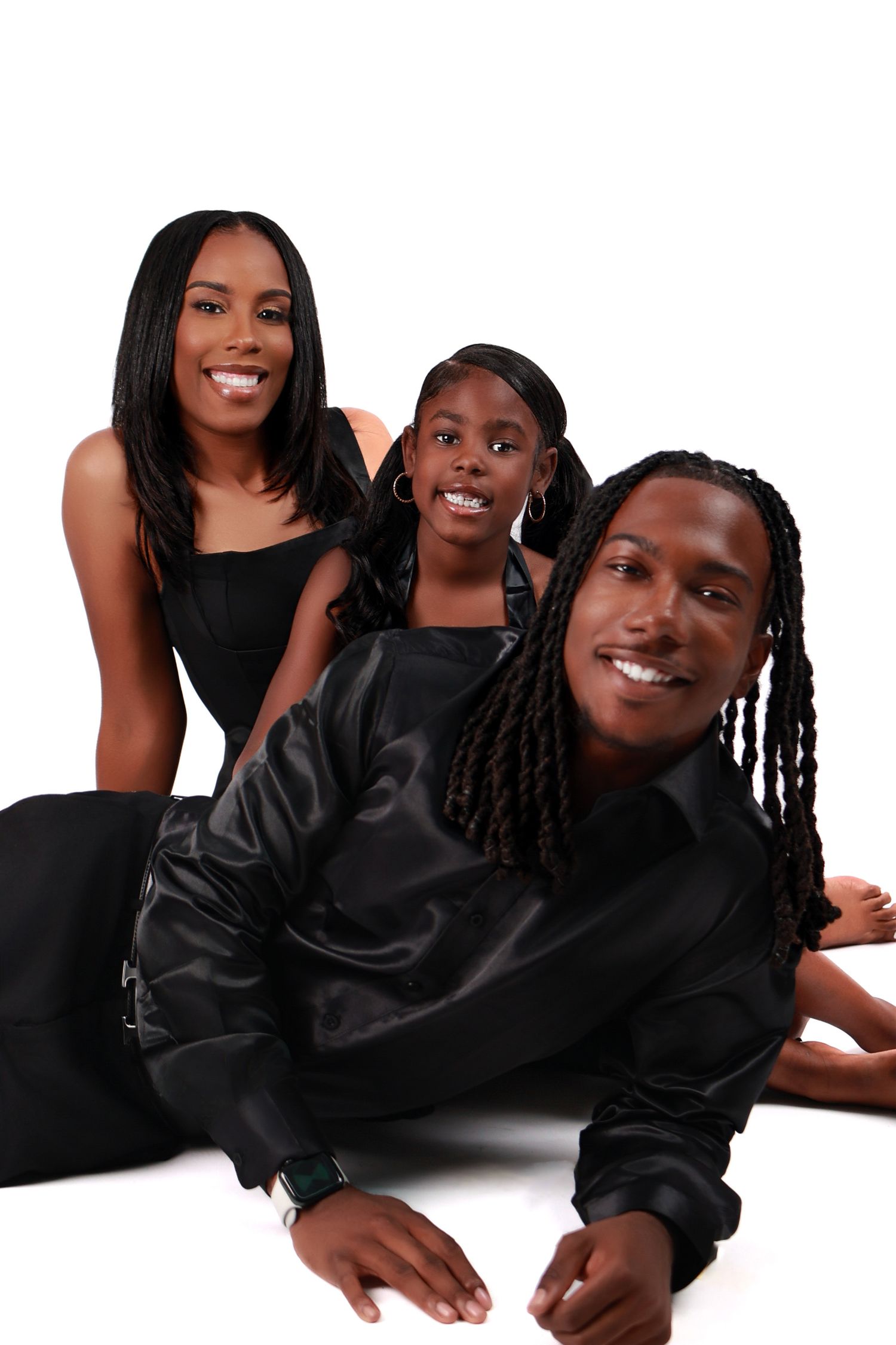Black family of three in matching black outfits pose together on a white studio floor smiling.
