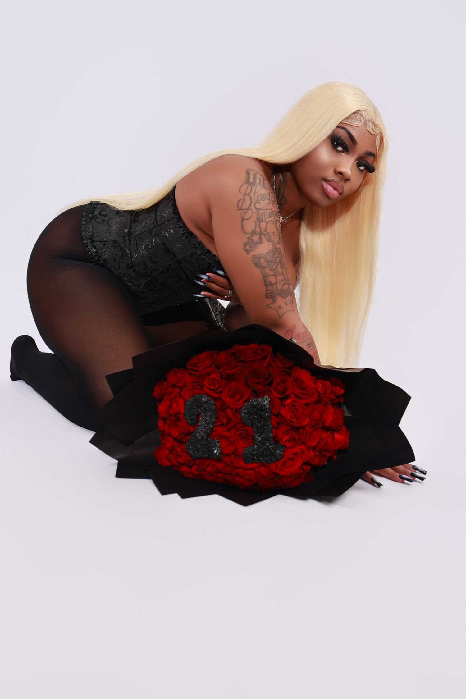 Woman with blonde hair and tattoos posing with large red rose bouquet on white background.