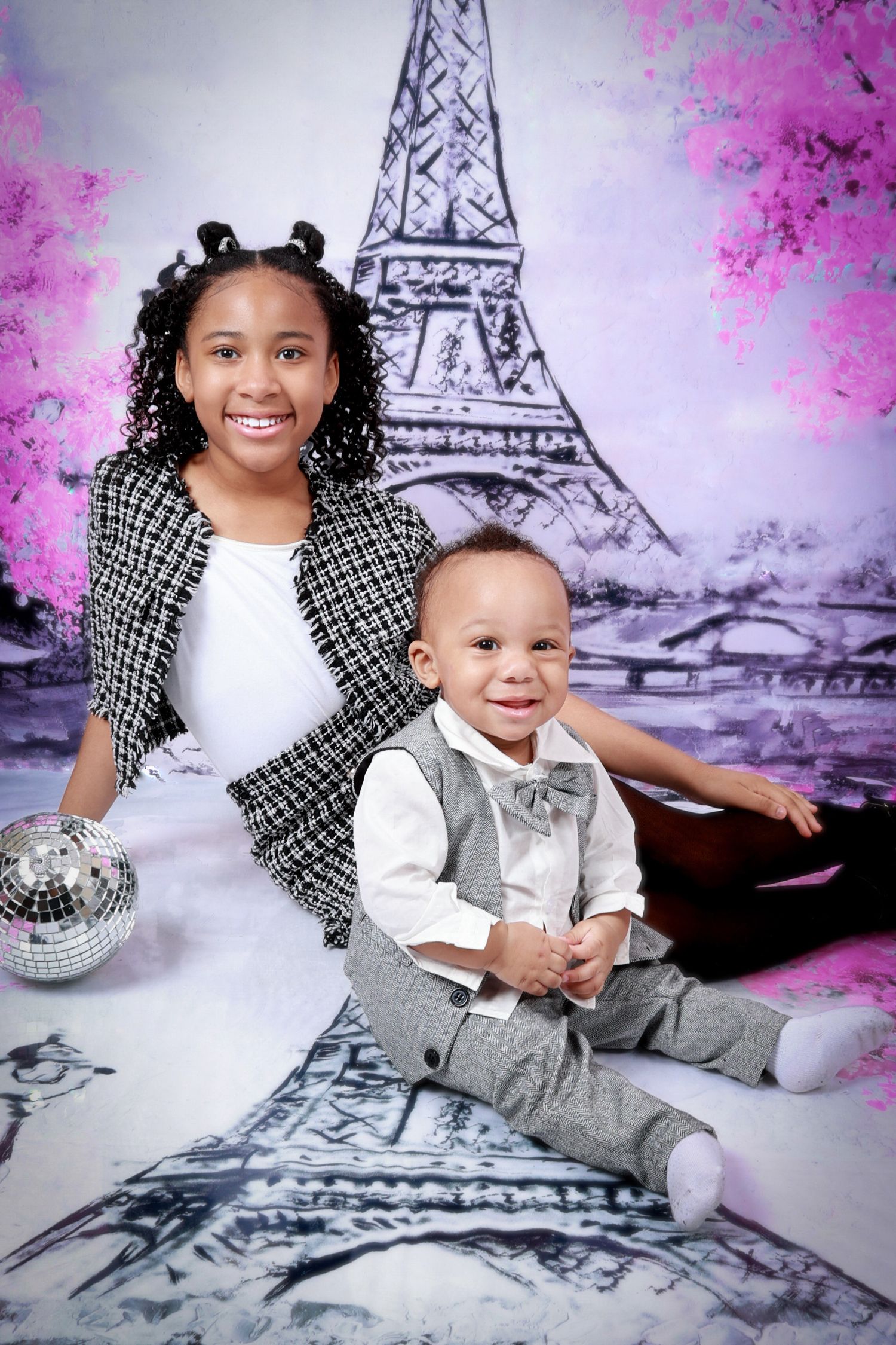 Two children pose in front of an Eiffel Tower backdrop with pink trees in a Paris-themed photo shoot.