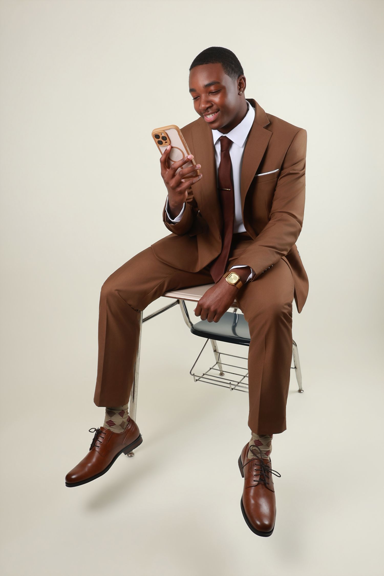 A well-dressed man in a brown suit sits on a chair while smiling at his smartphone.