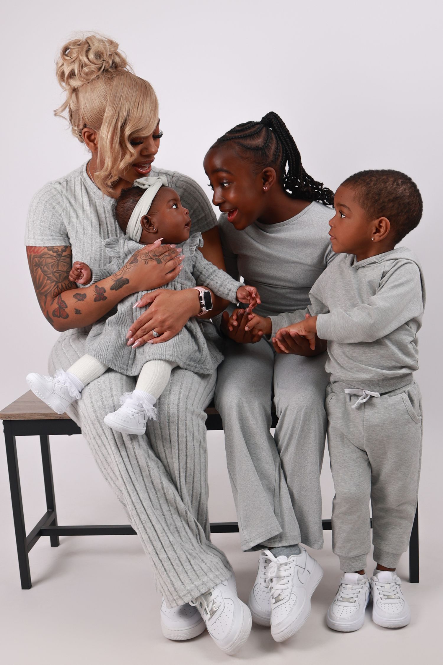 A mother sits on a bench with three young children all wearing matching gray outfits in a studio photo.