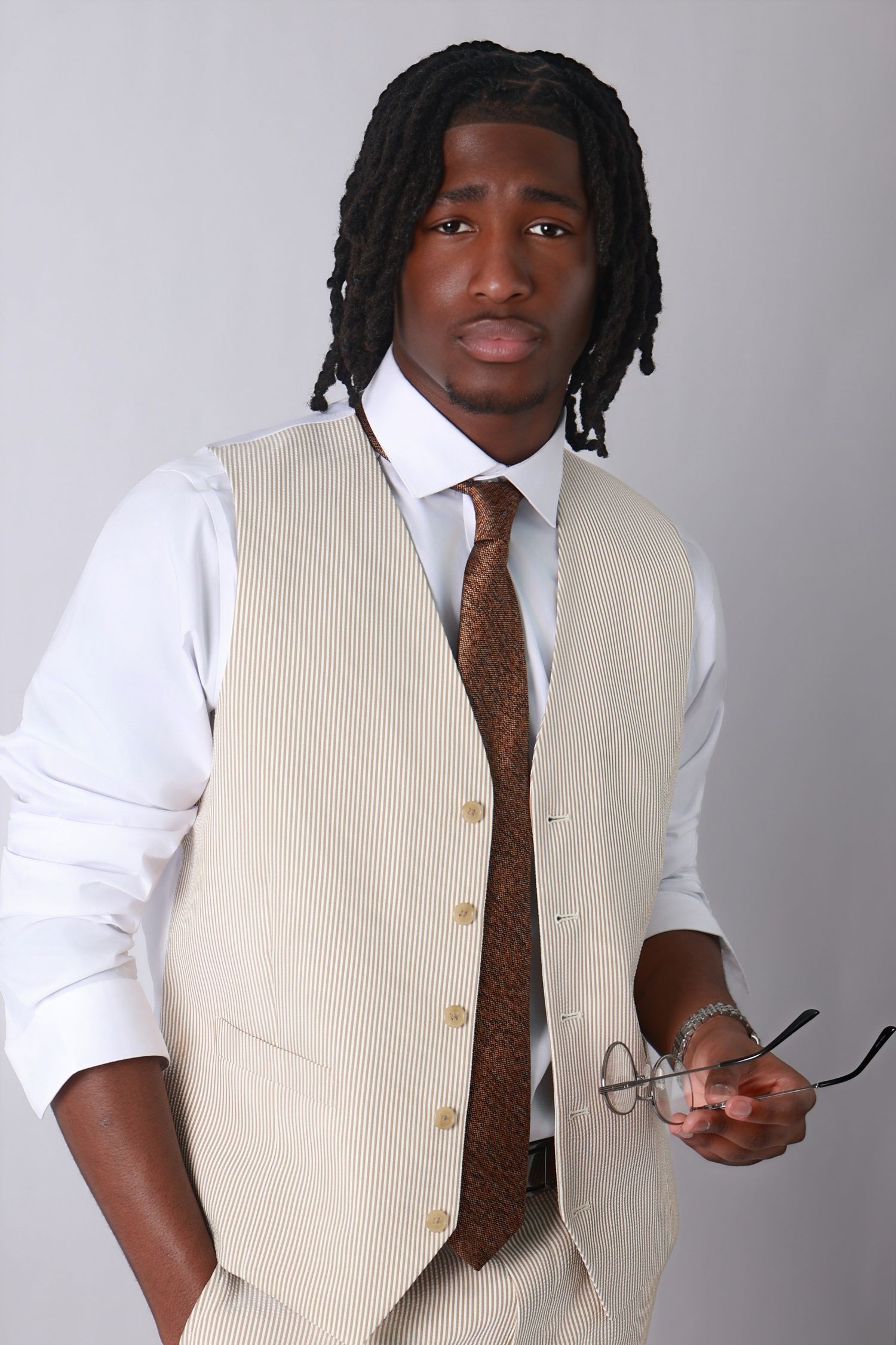 Young Black man with dreadlocks wearing a beige vest and tie holds glasses in a professional studio portrait.