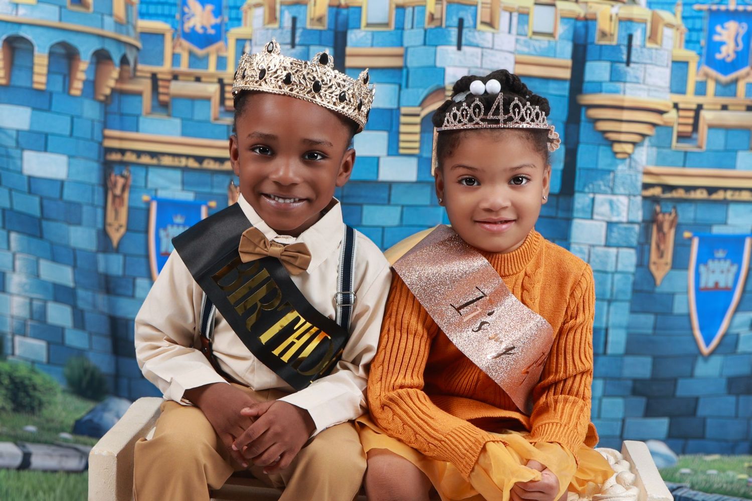 Two young children (twins) wearing royal crowns and sashes posing in front of castle backdrop.