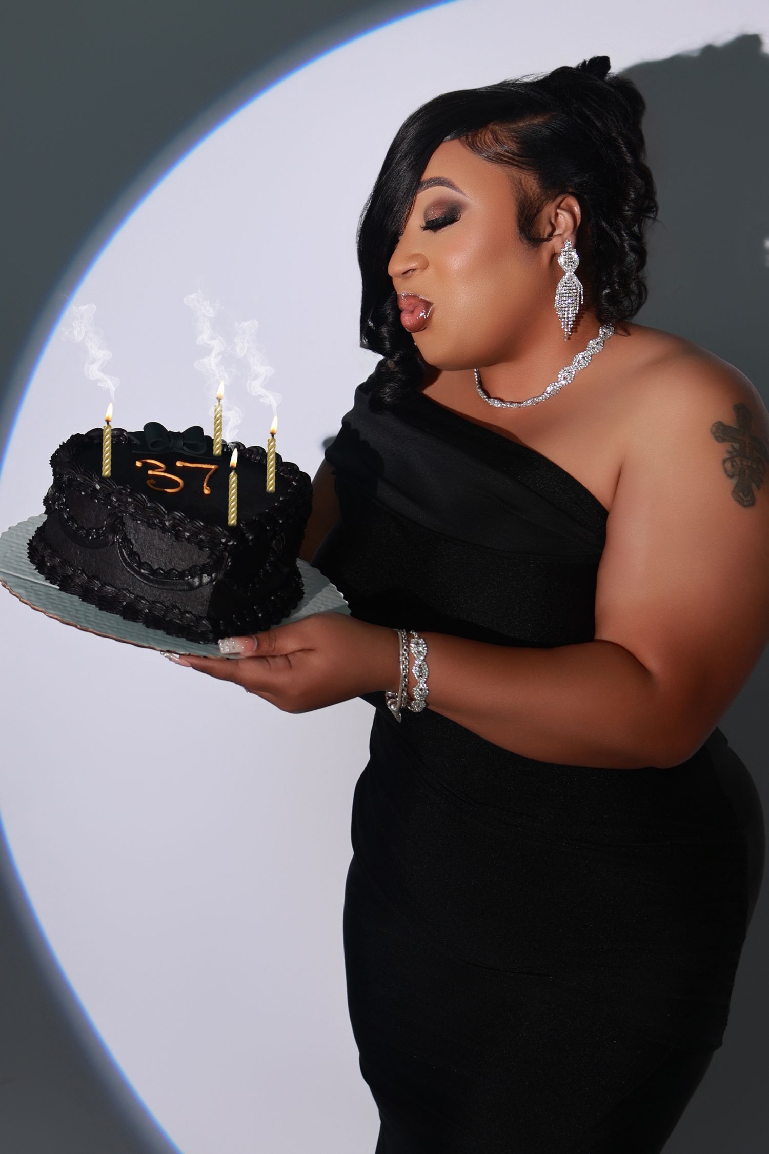 Woman in black one-shoulder dress blows out candles on a dark birthday cake in a dramatic spotlight portrait.
