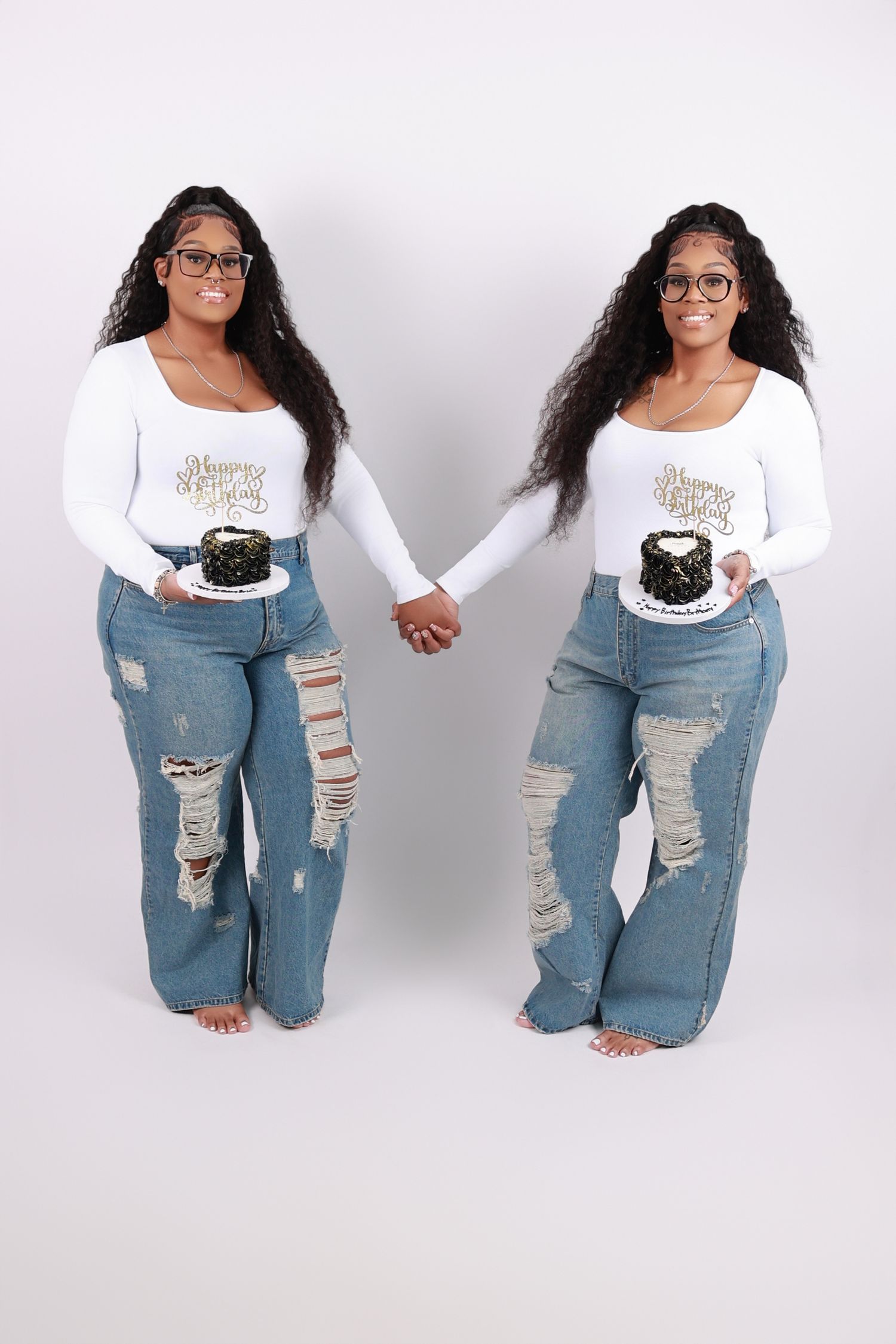 Twin sisters in matching white tops and ripped jeans holding birthday cakes hand in hand.